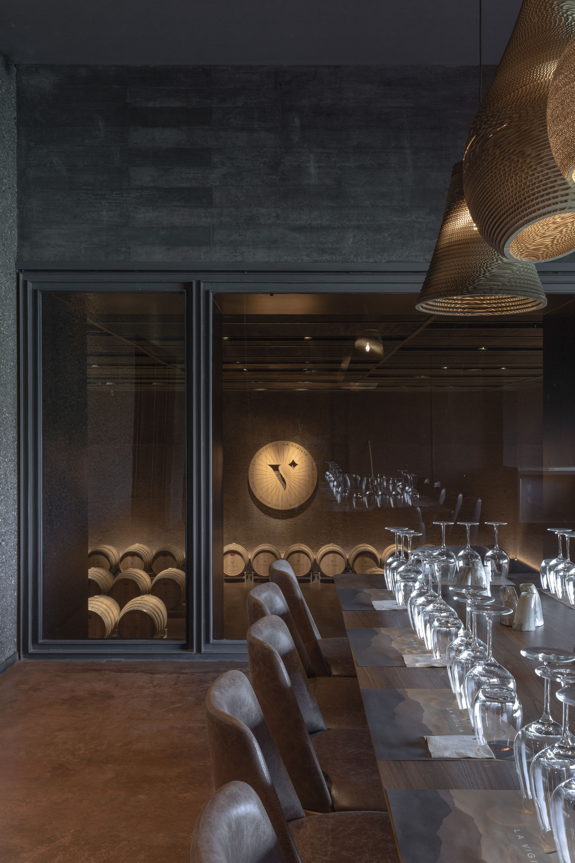 Long communal table with leather chairs and upturned wine glasses, facing a glazed wall that reveals stacked barrels in warm light