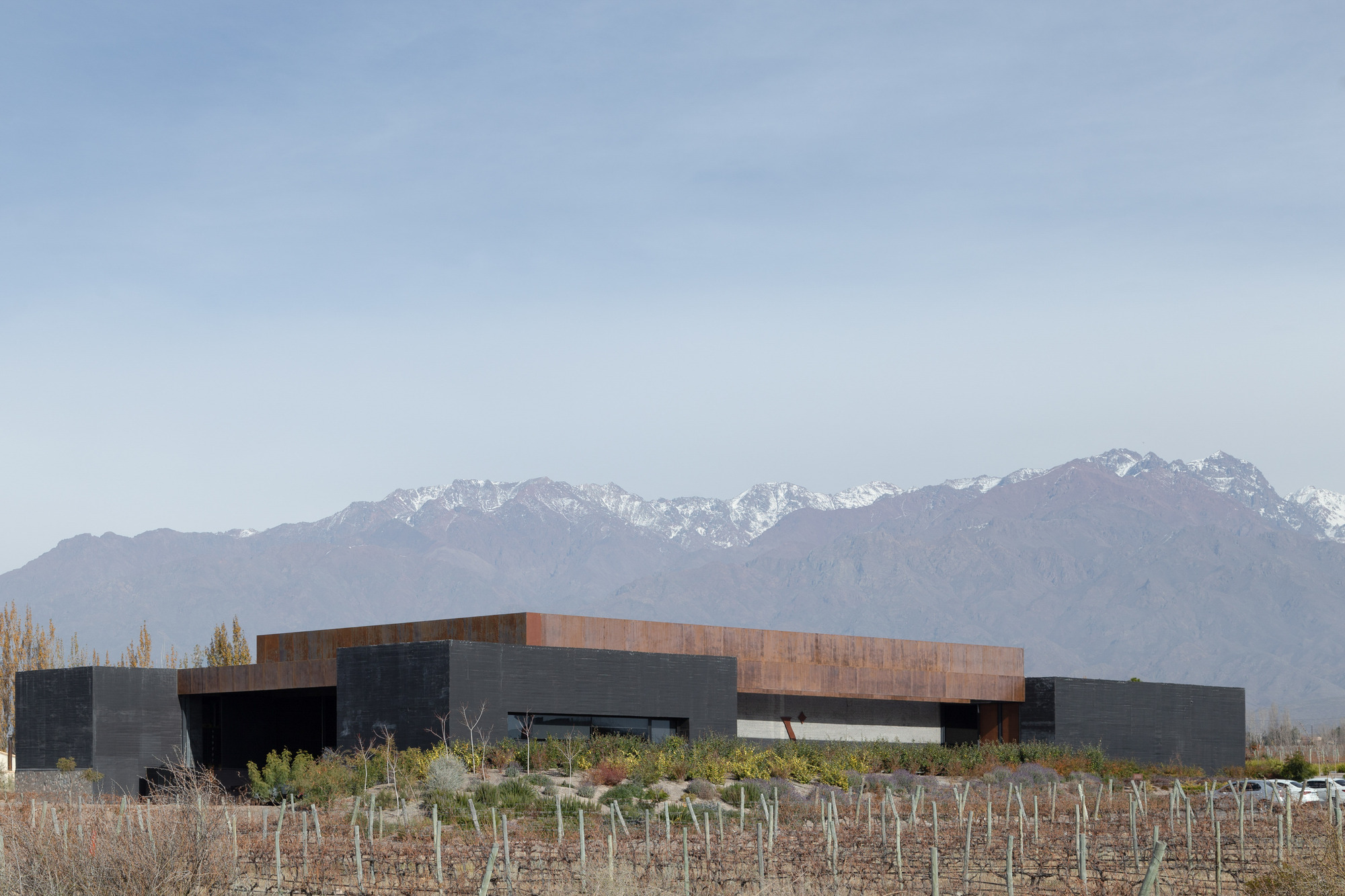 Low dark concrete volumes topped by a weathering steel band, set behind bare vines with snowy mountains beyond