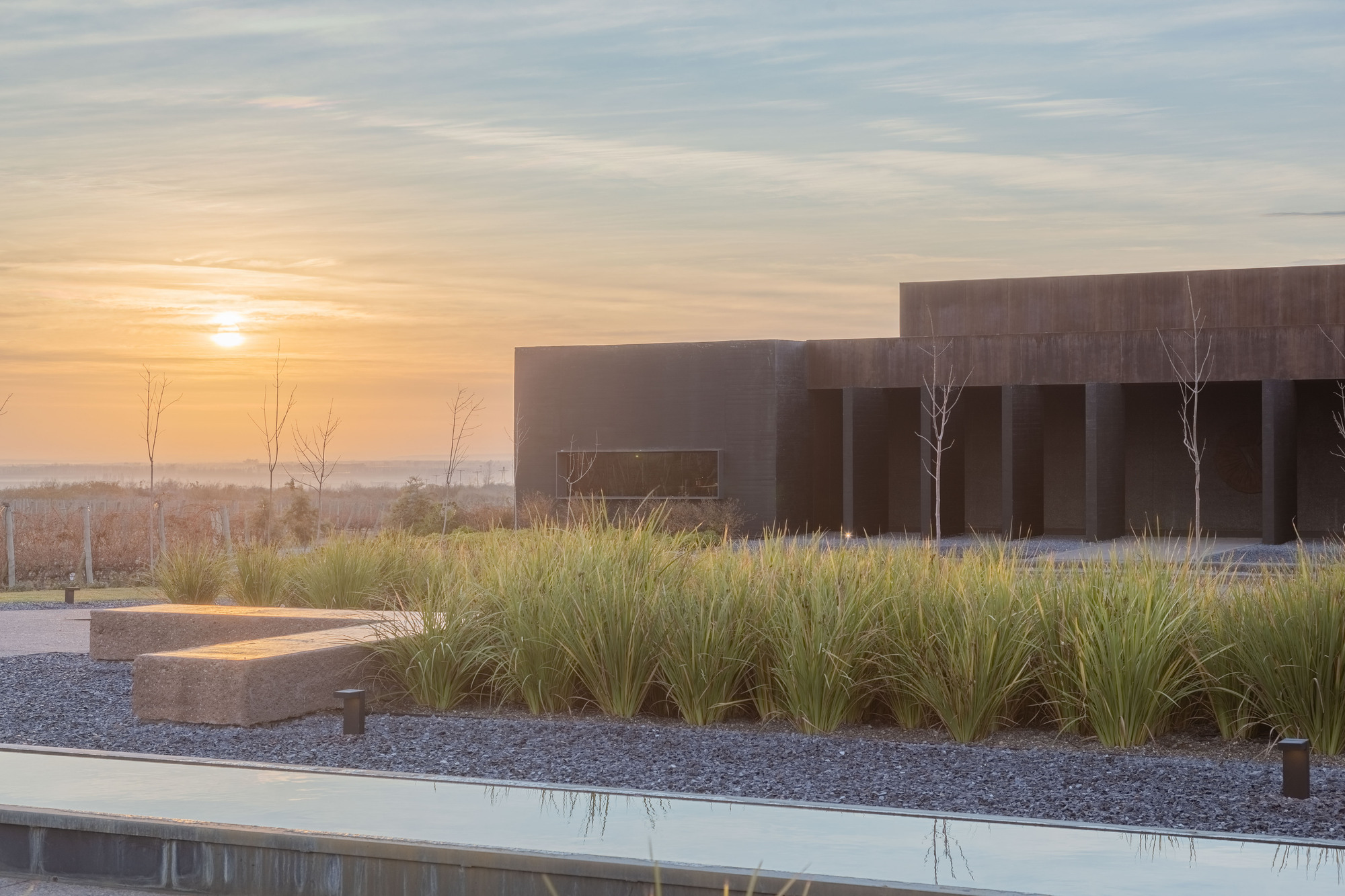 Sunrise over a gravel bed of tall grasses and a still water channel, with a colonnaded porch beyond