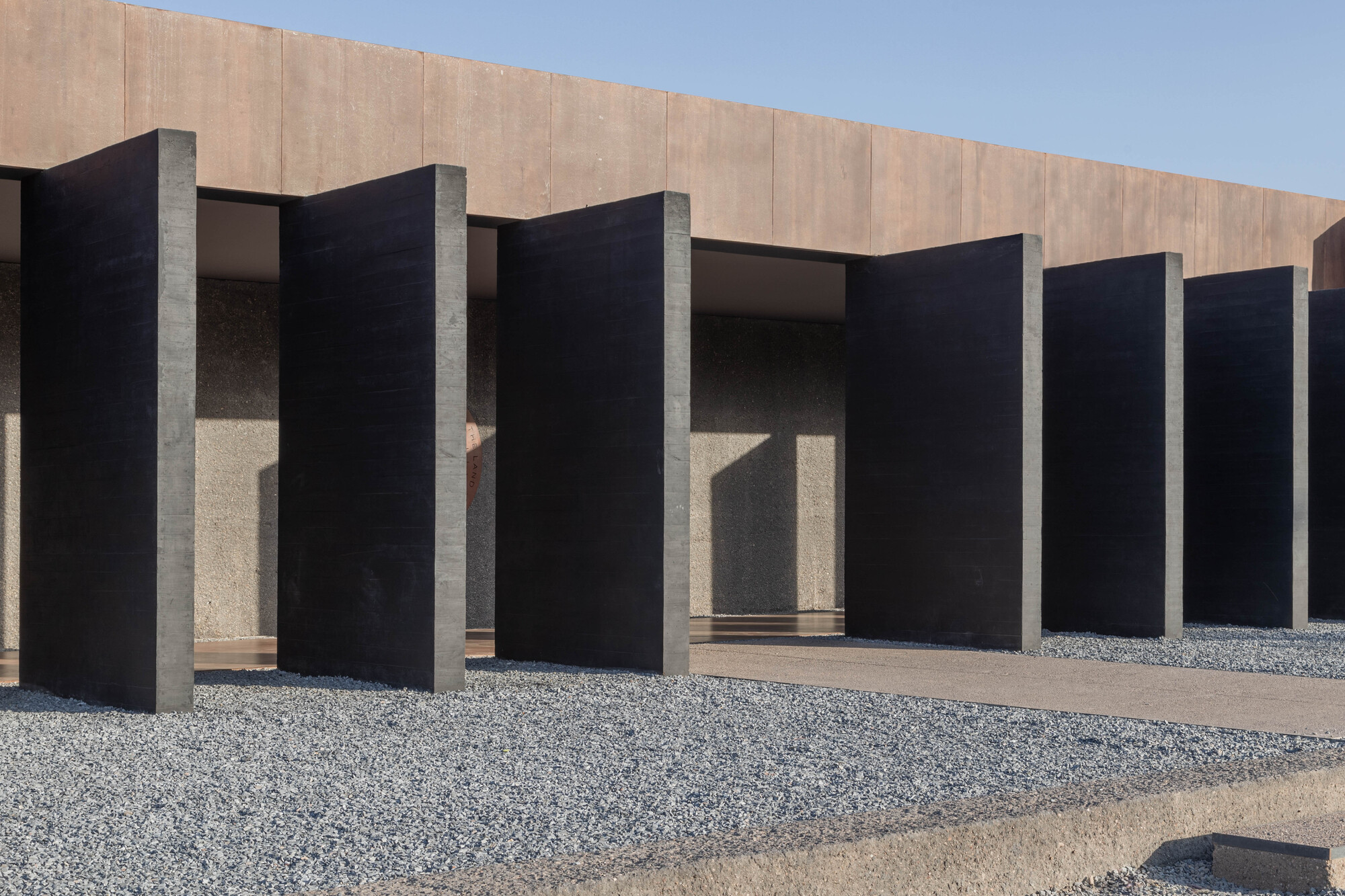 Row of tall black concrete fins standing over gravel in front of a weathering steel facade under clear blue sky