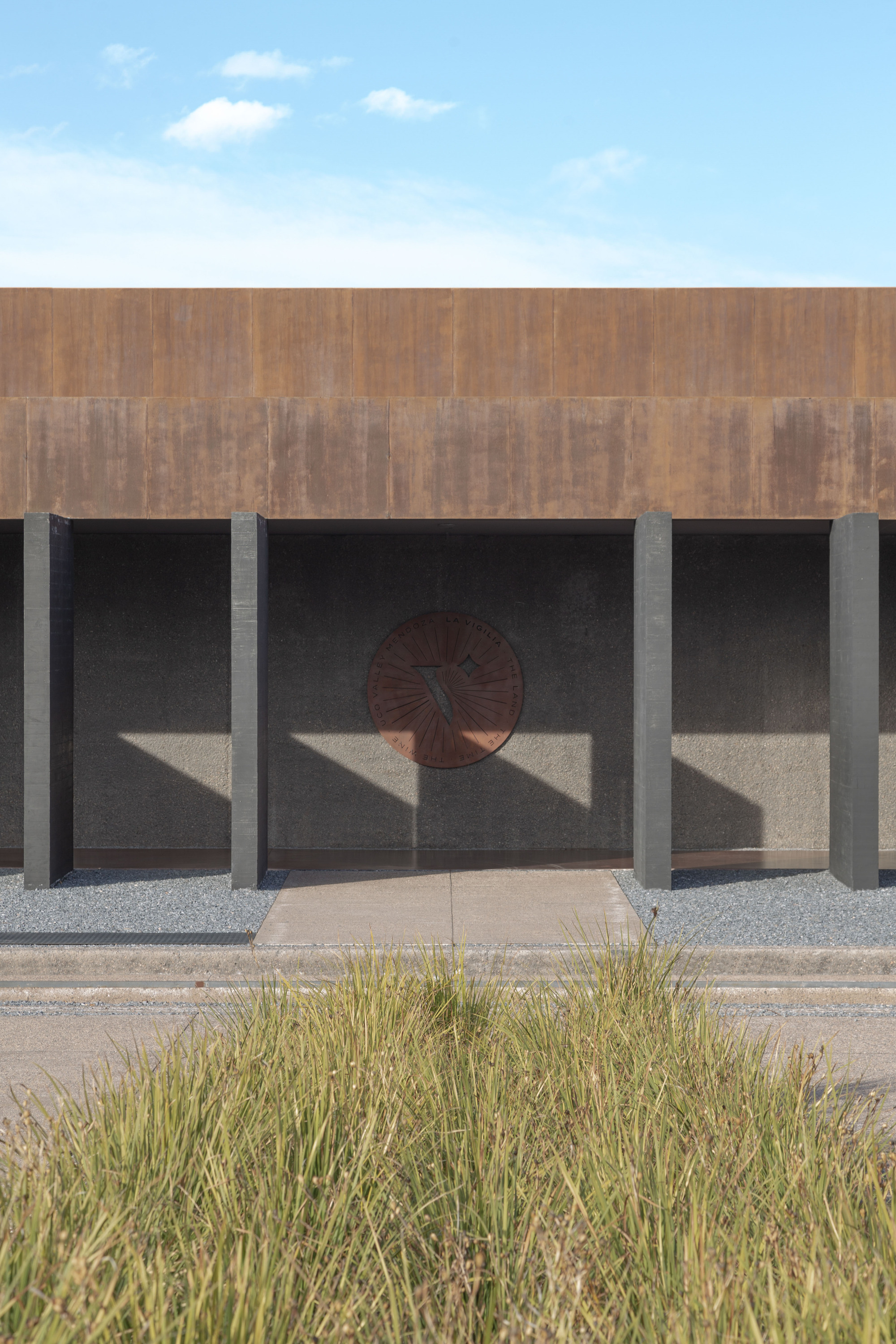 Weathering steel facade above slim dark columns and a circular rusted emblem, with tall grass in the foreground and light cloud