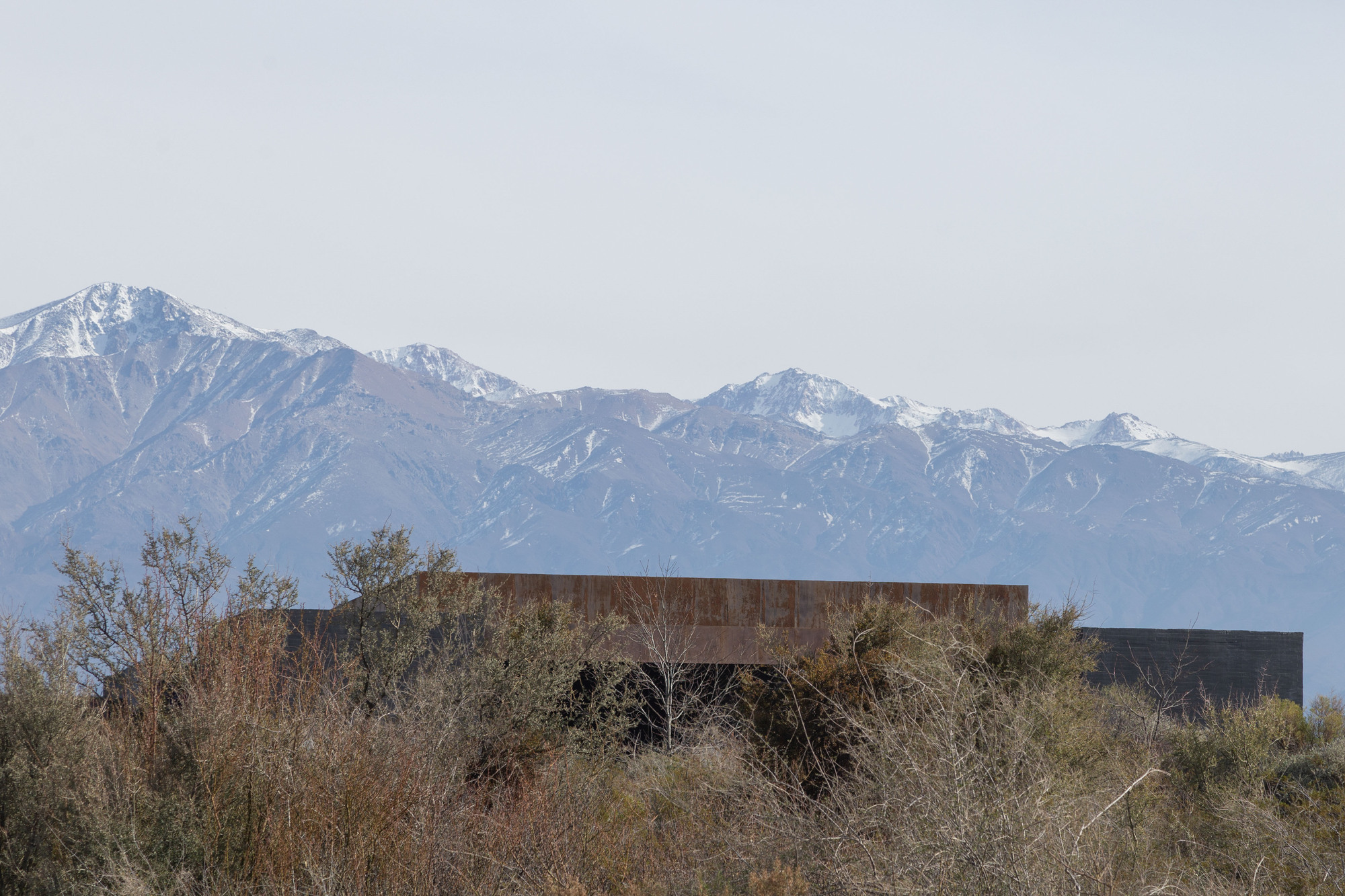 Low weathering steel volume half hidden by dry scrub, with snow-capped mountains rising behind in hazy light
