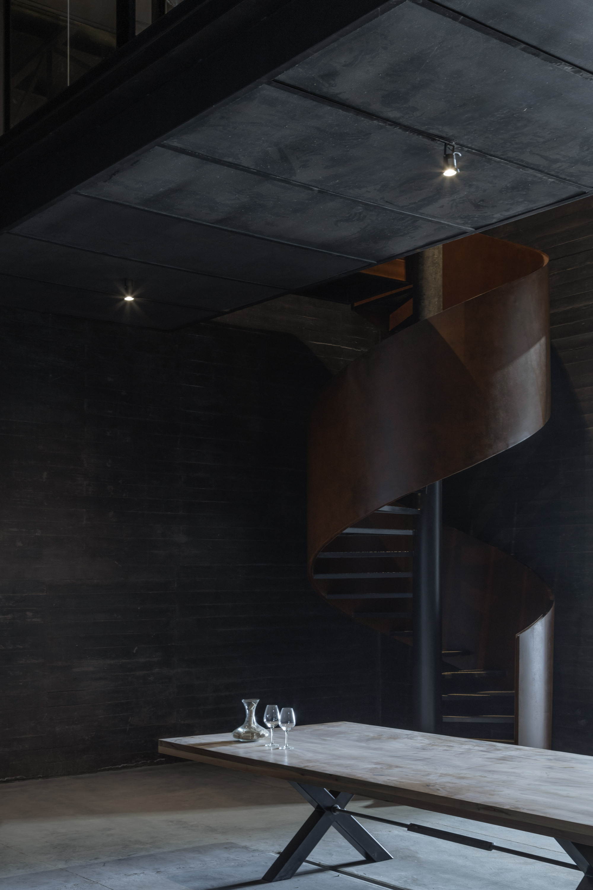 Spiral staircase of rusted steel beside a dark brick wall, with a timber table, carafe and wine glasses in the foreground