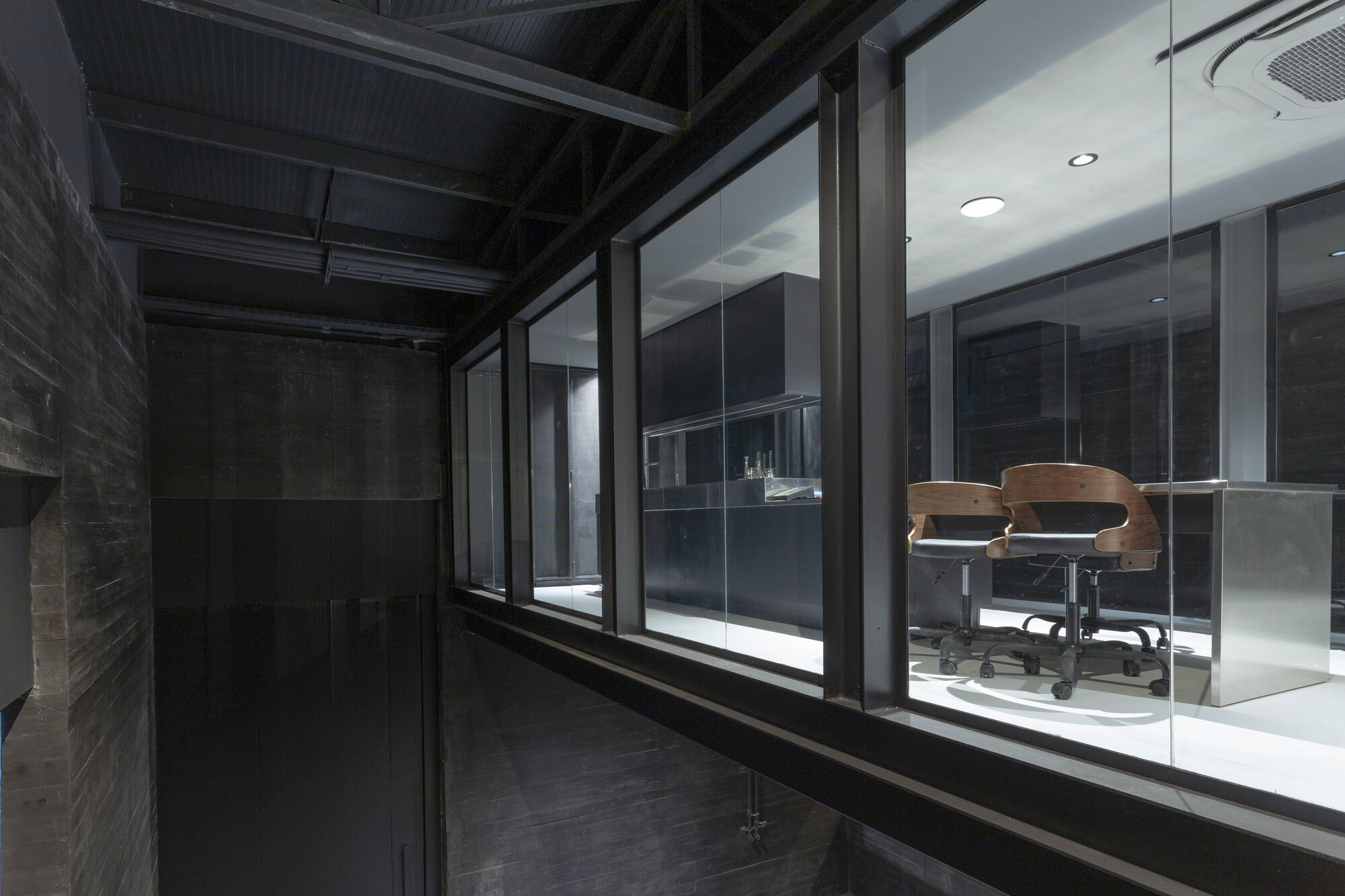 Steel-framed glazing looking into a lit tasting room with wooden swivel chairs, seen from a dark board-formed concrete corridor at night