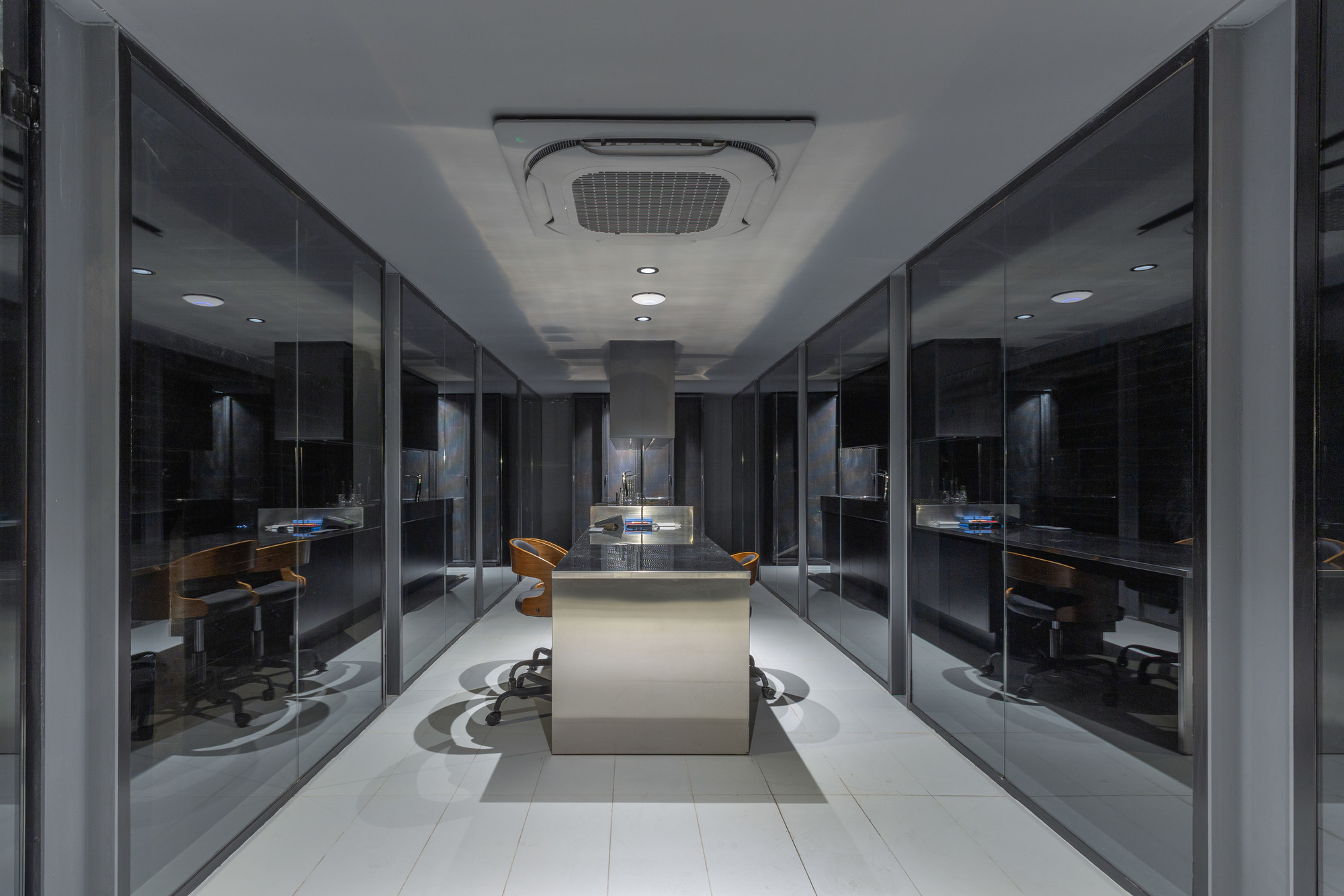 Central stainless steel island table between glass-walled rooms, with orange chairs and a ceiling cassette unit under cool evening light