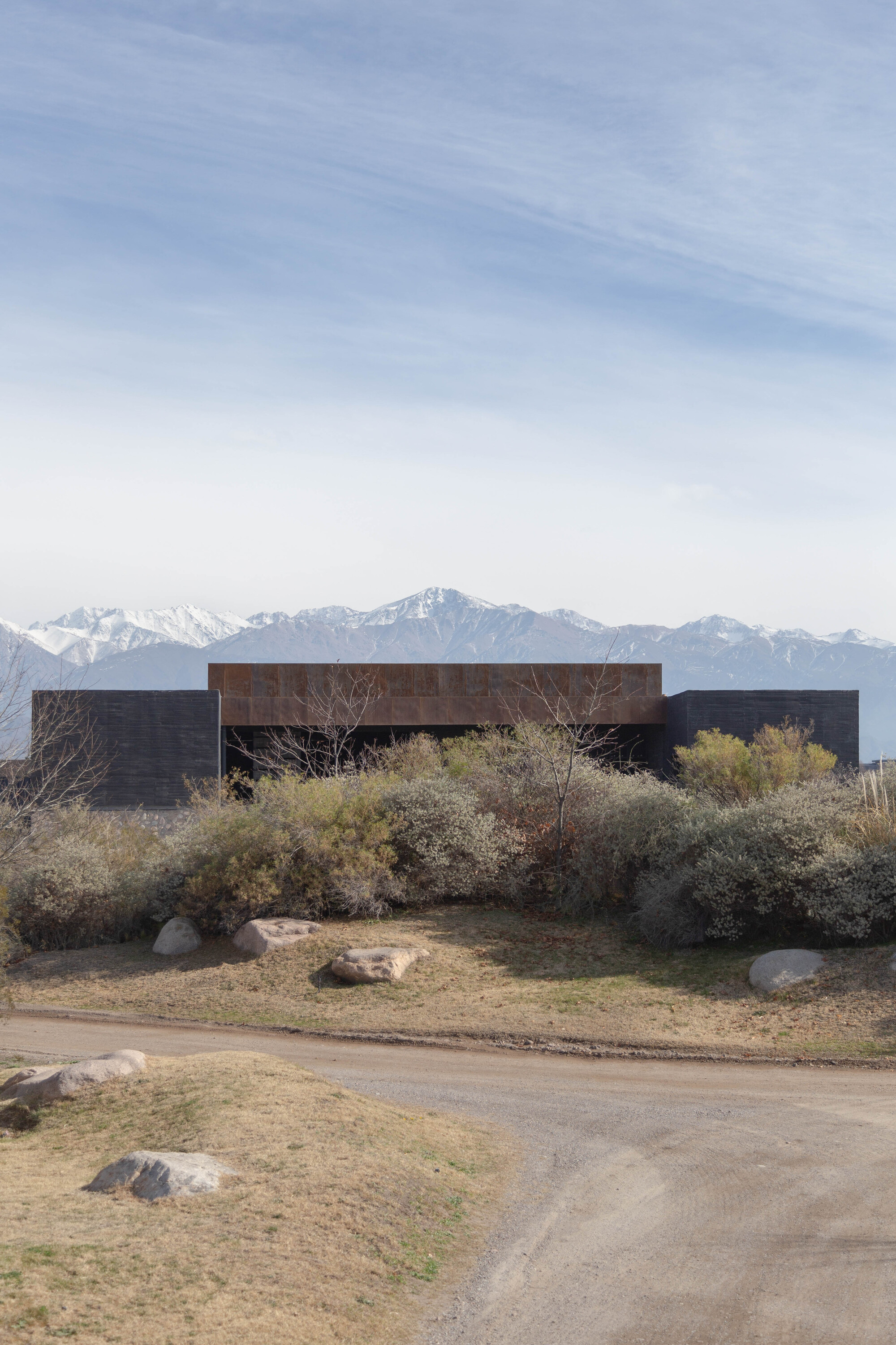 Low weathered steel and dark concrete volume behind dry shrubs and boulders, with snowcapped mountains under a hazy sky
