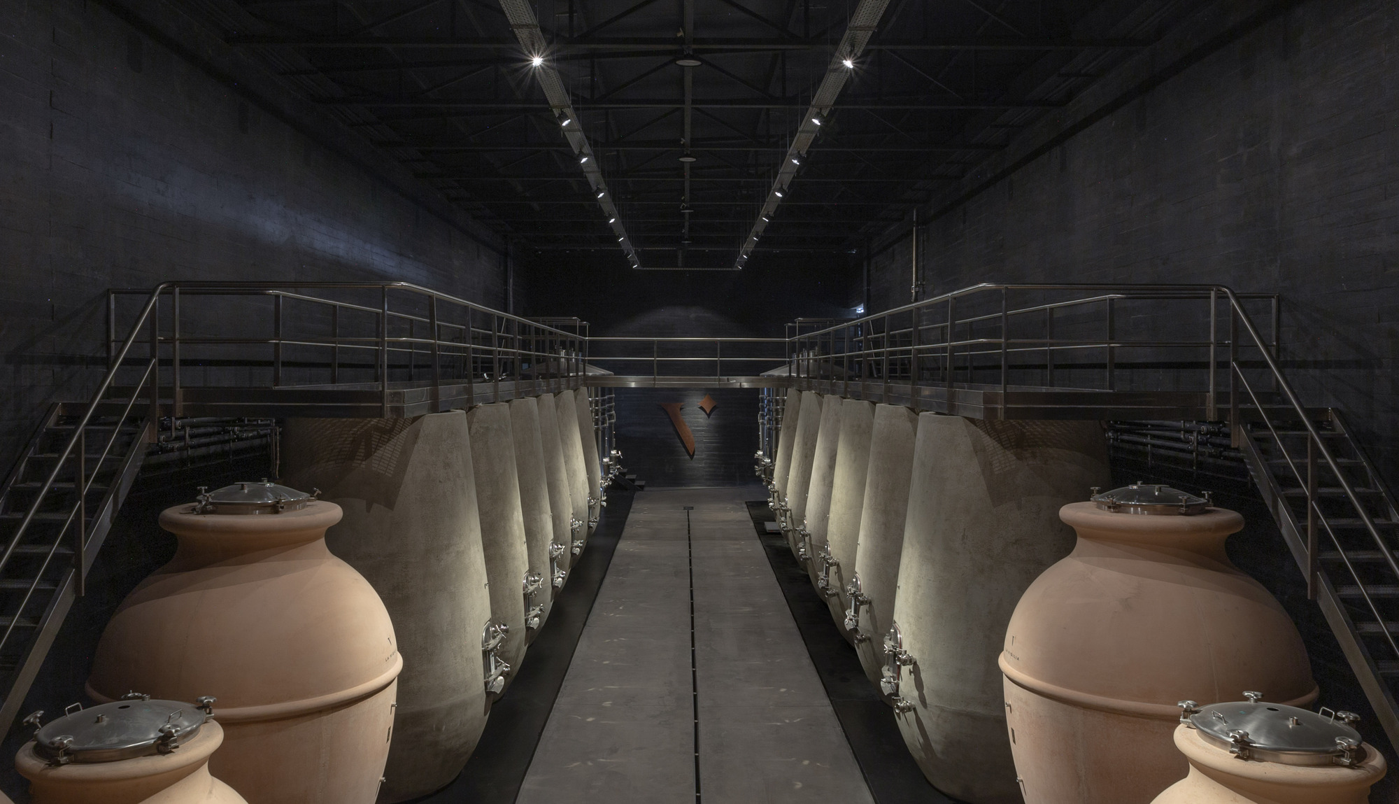 Row of large clay fermentation vessels beside steel catwalks and stairs in a dark concrete hall lit by track spotlights