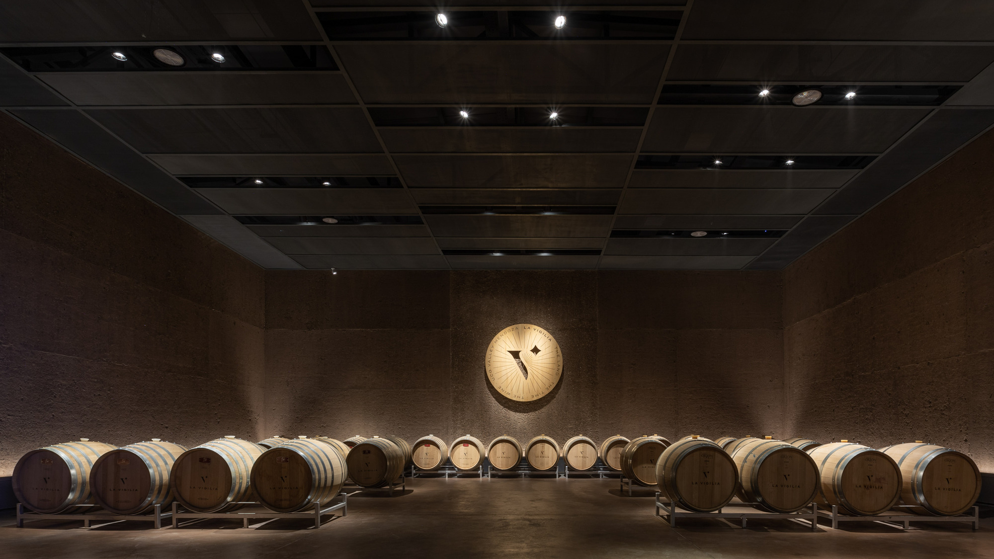 Oak barrels in rows facing a round emblem on a rammed earth wall, with a dark coffered ceiling above