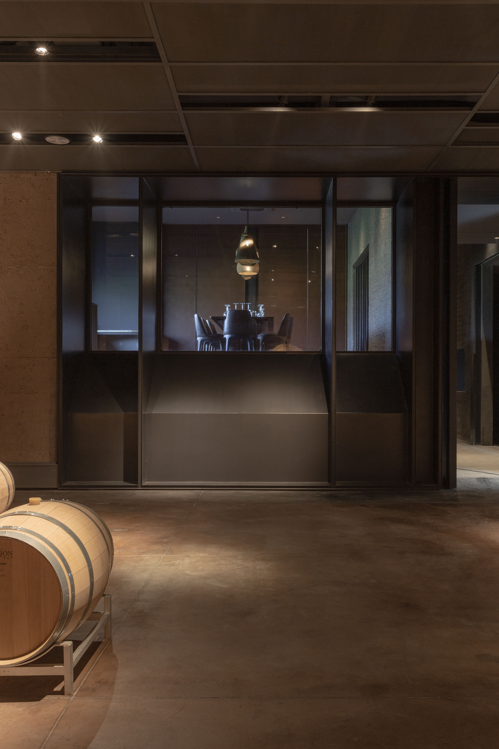 Oak wine barrel on a steel rack before a dark metal-framed glazed partition, with a dining table glowing under a pendant lamp