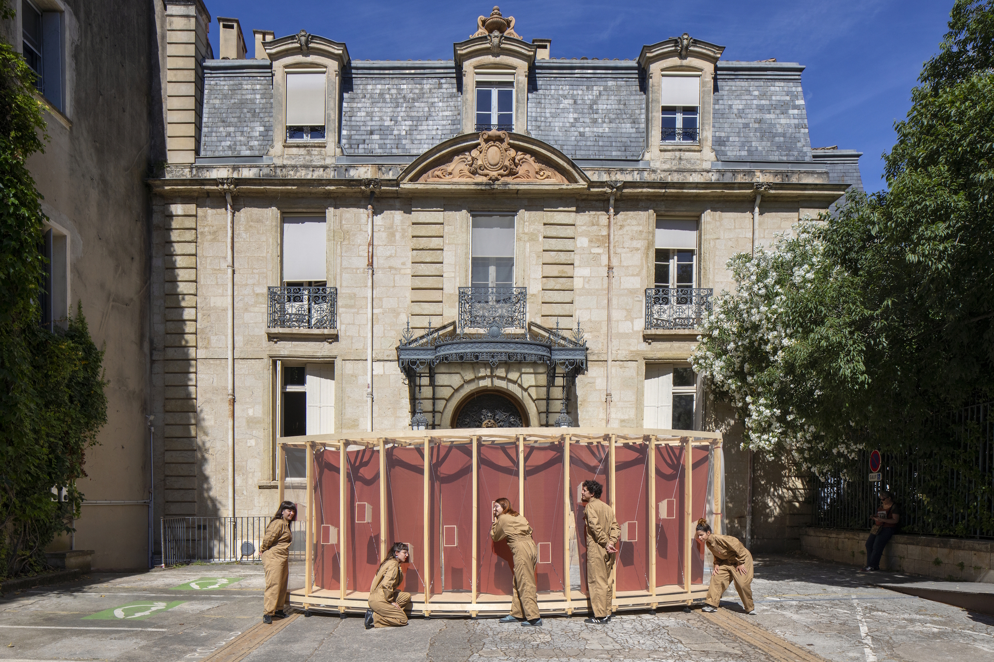 Curved timber-framed pavilion with red fabric panels in a courtyard, with people in tan overalls pushing it beneath a stone mansard facade