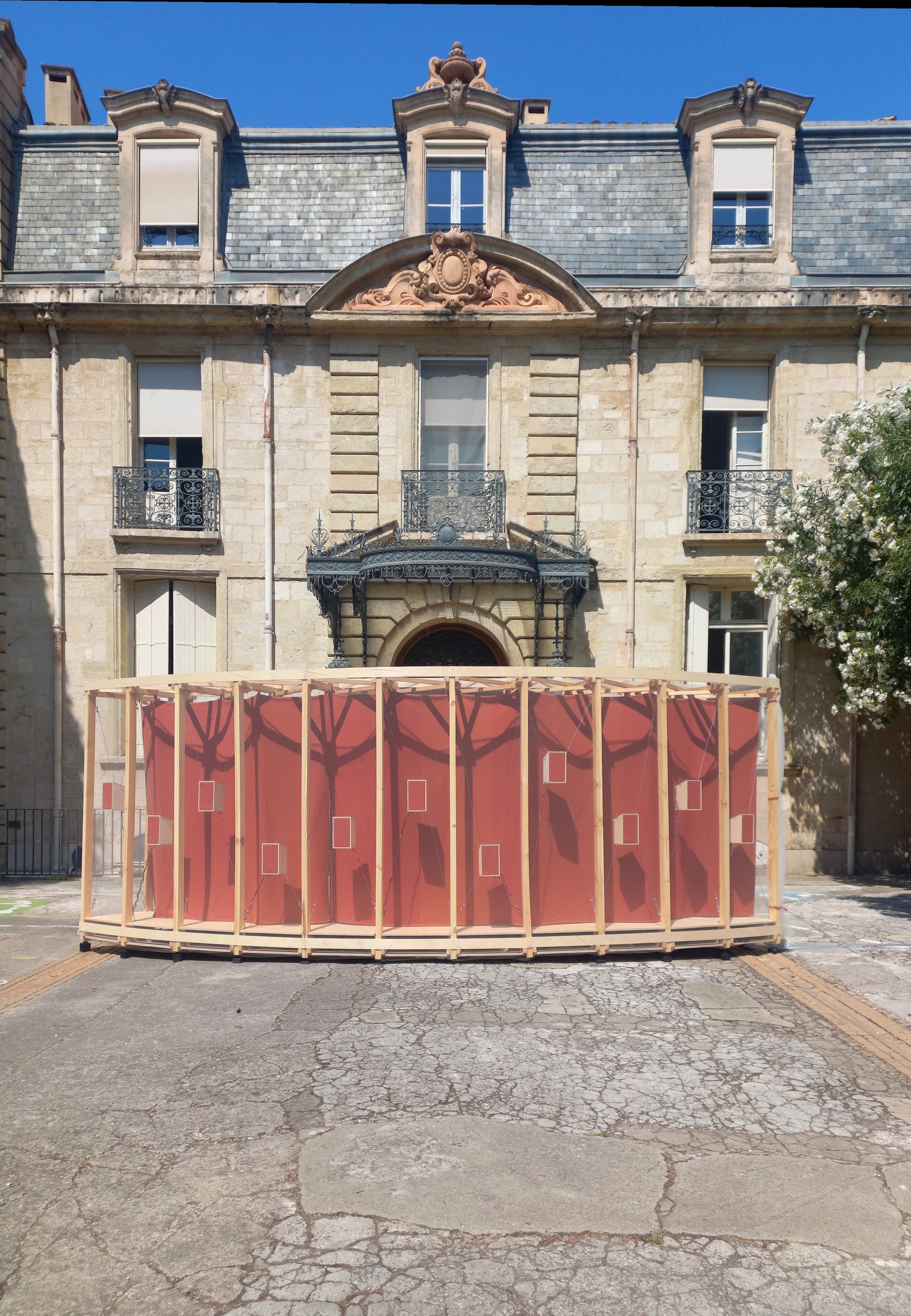 Frontal view of the red-lined timber pavilion standing on cracked paving in front of a stone facade under blue sky
