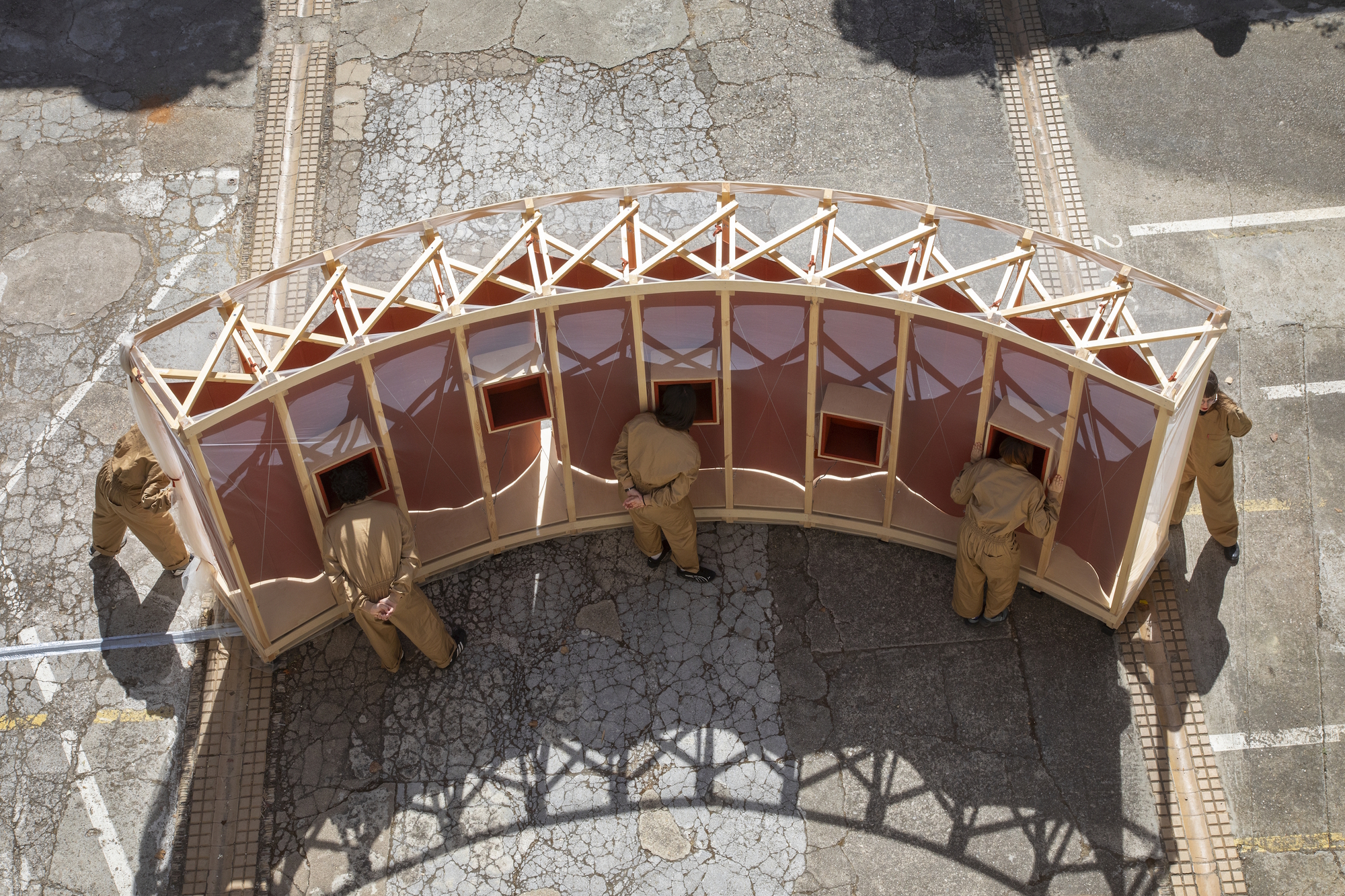 Top-down view of the arced pavilion, with people in tan overalls leaning to its viewing boxes and a lattice roof casting shadows