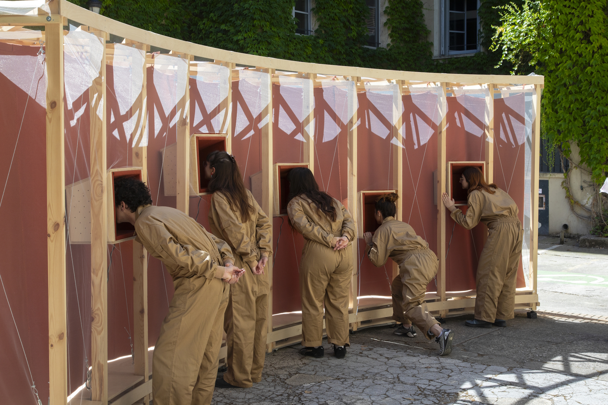 Row of people in tan overalls bending to peer into small wooden boxes set in the red curved wall, with ivy behind