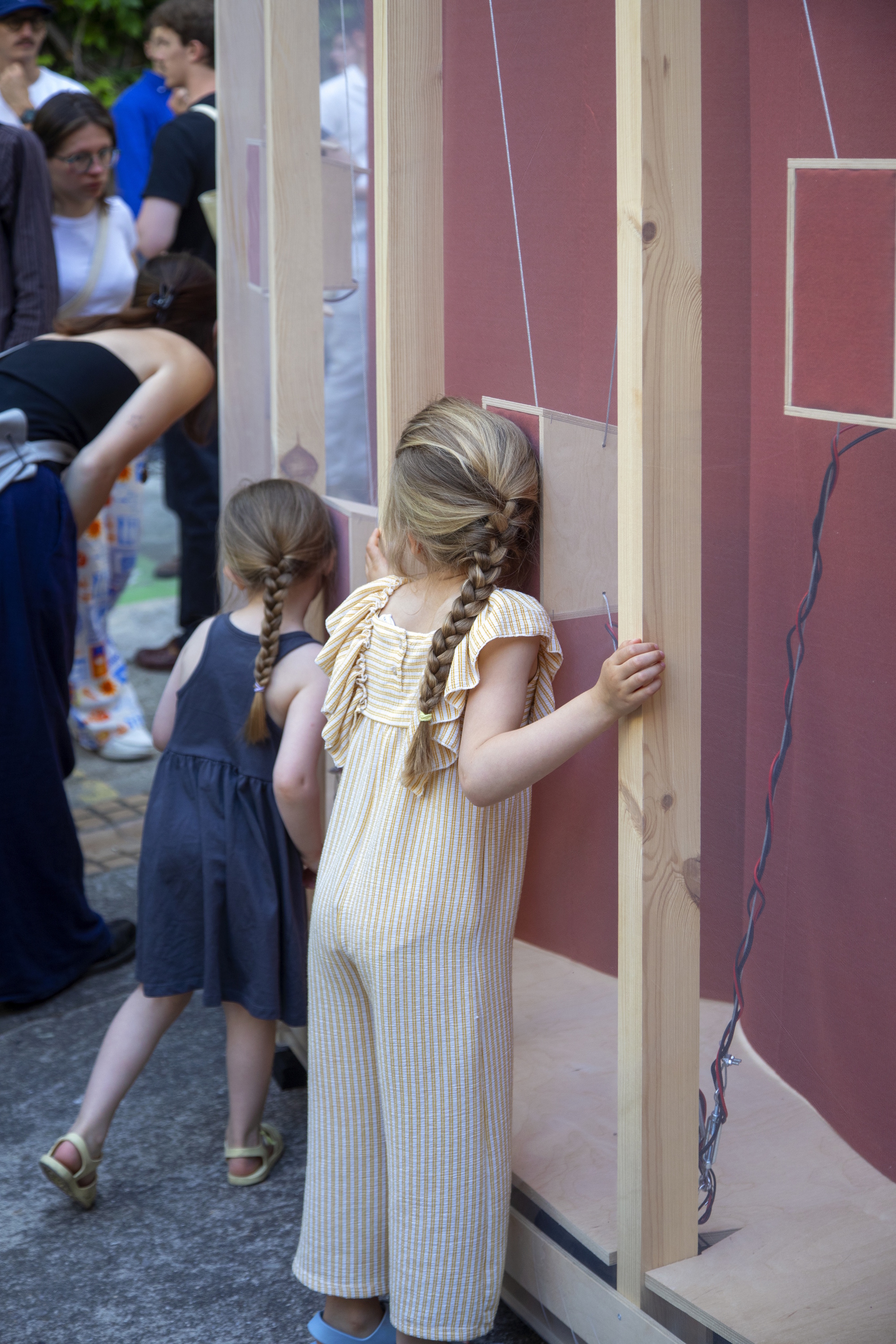 Girl with long braids pressing her face to the red panel beside a timber post, with a smaller child and onlookers behind