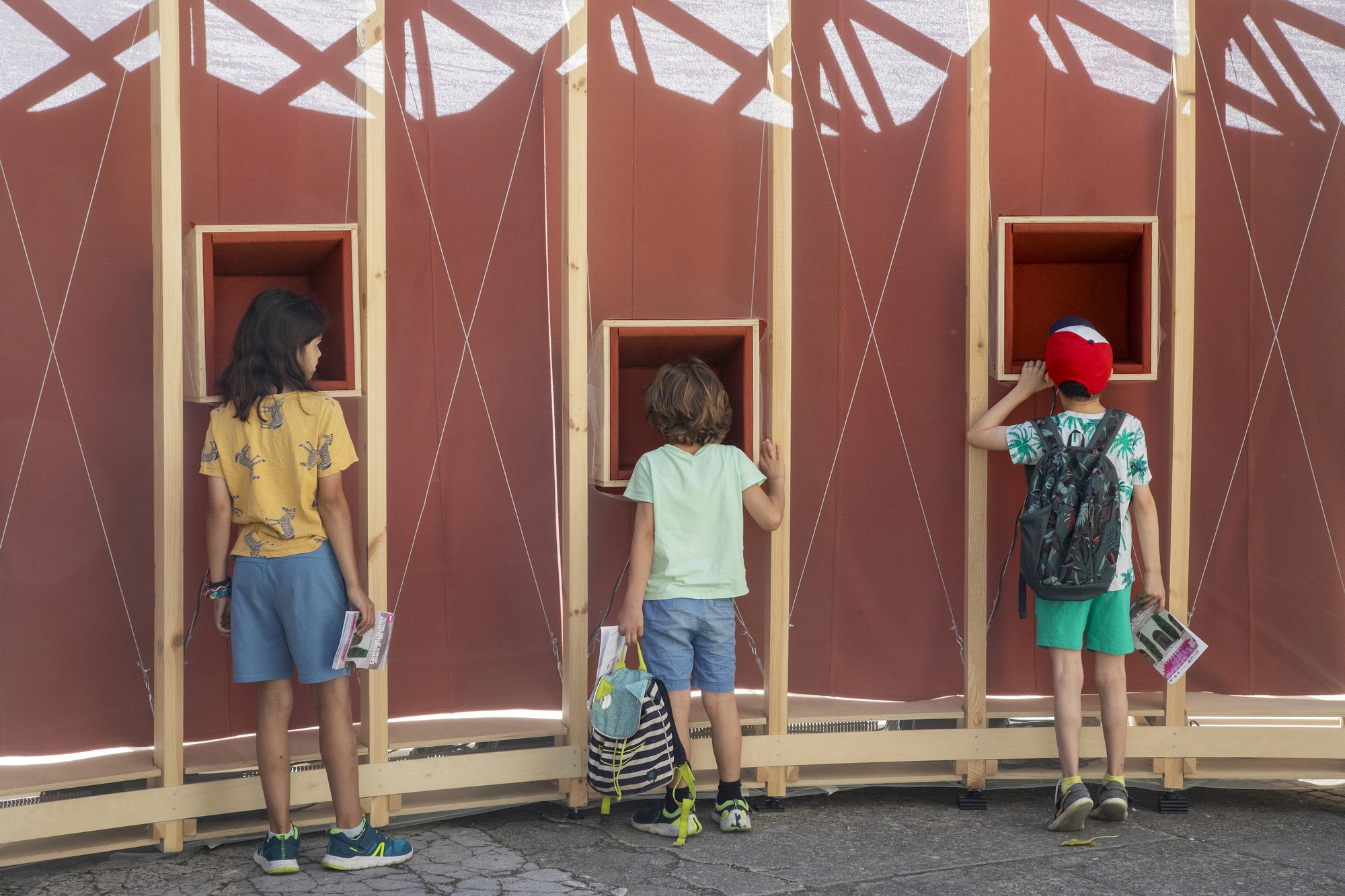 Three children in summer clothes looking into recessed wooden boxes in the red fabric wall, with sunlit rope lines across it