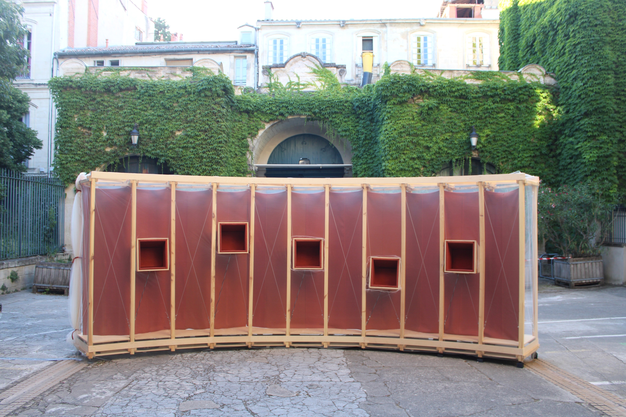 Curved timber-framed pavilion with rust-red fabric panels and square openings, set in a courtyard before an ivy-covered wall