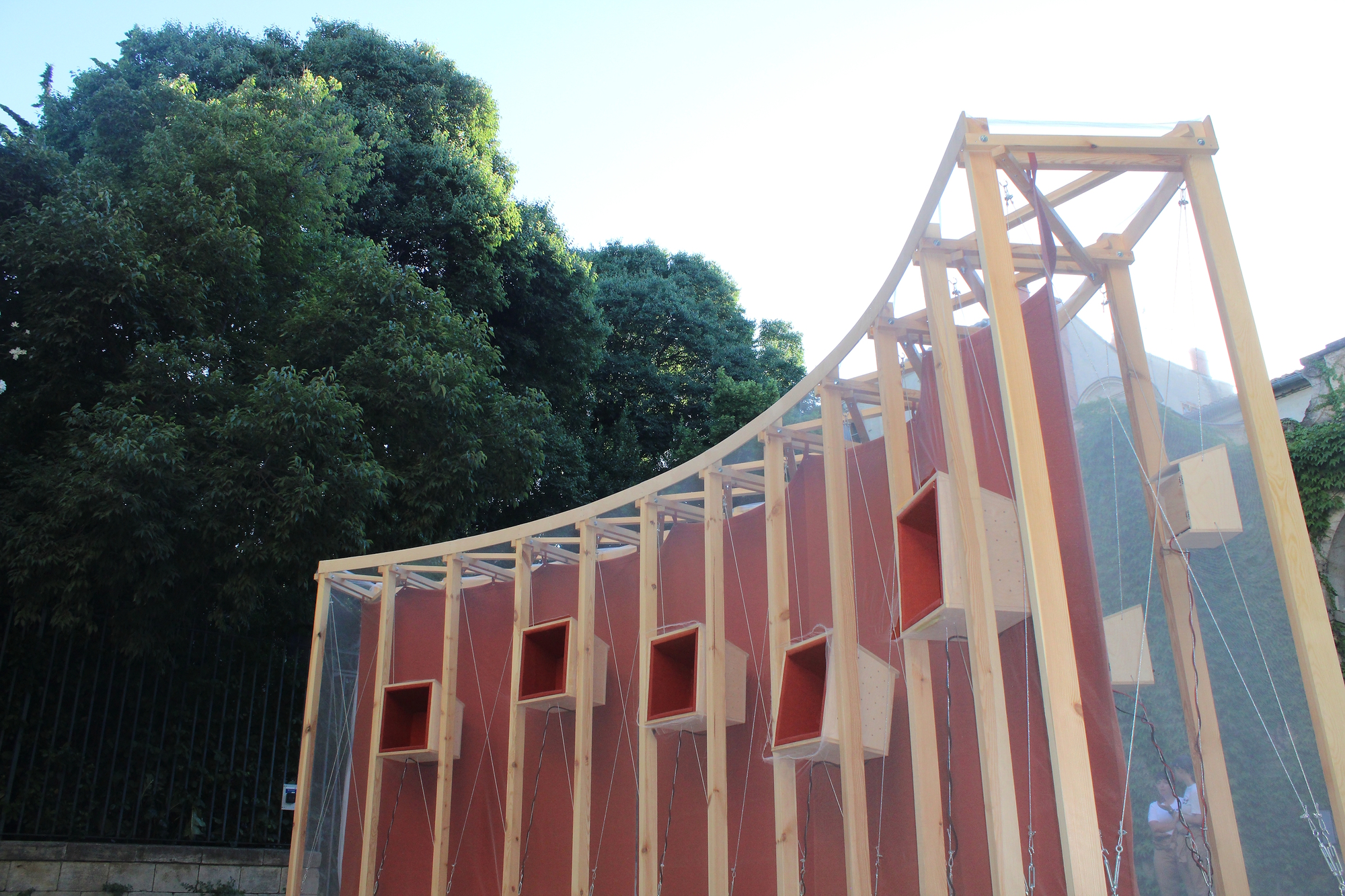 Side view of the curved timber frame with red panels and boxy wooden speaker cases, beside a dark tree in evening light