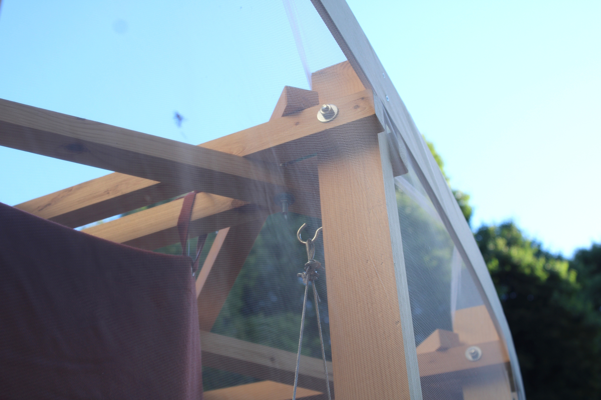 Close-up of a bolted timber joint and hanging wire hook seen through translucent mesh against a blue sky