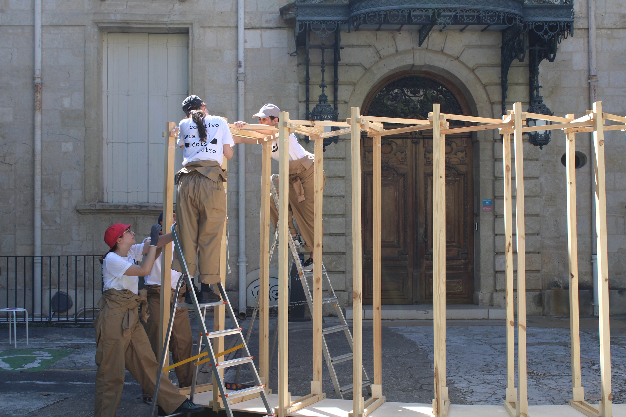 Builders in tan overalls on stepladders assembling the upright timber frame in front of a stone facade with an arched door