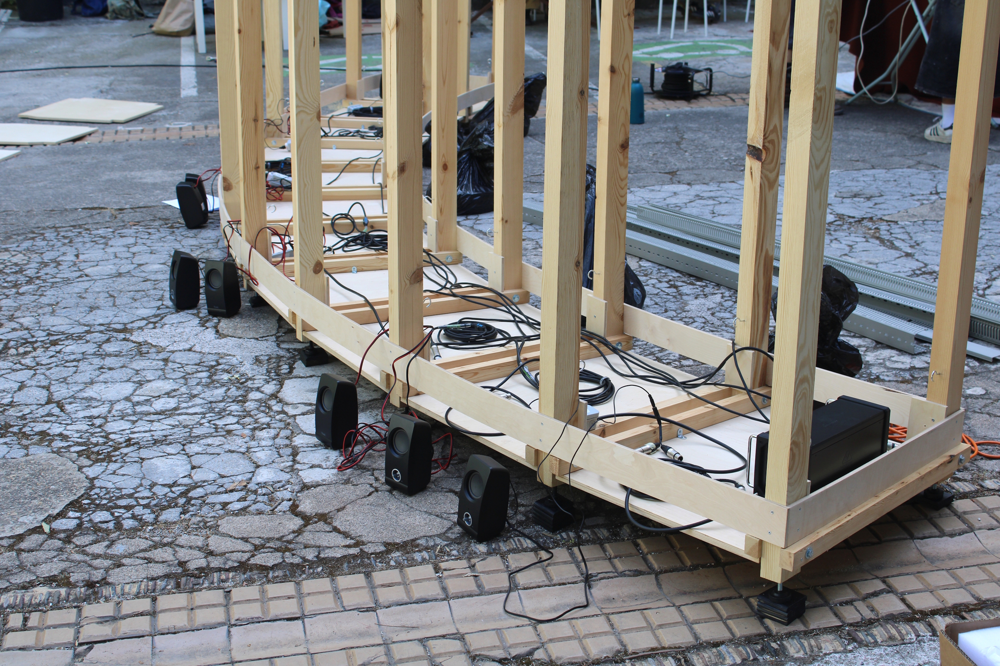 Timber base frame on cracked paving, with small black speakers and tangled cables laid out along the floor