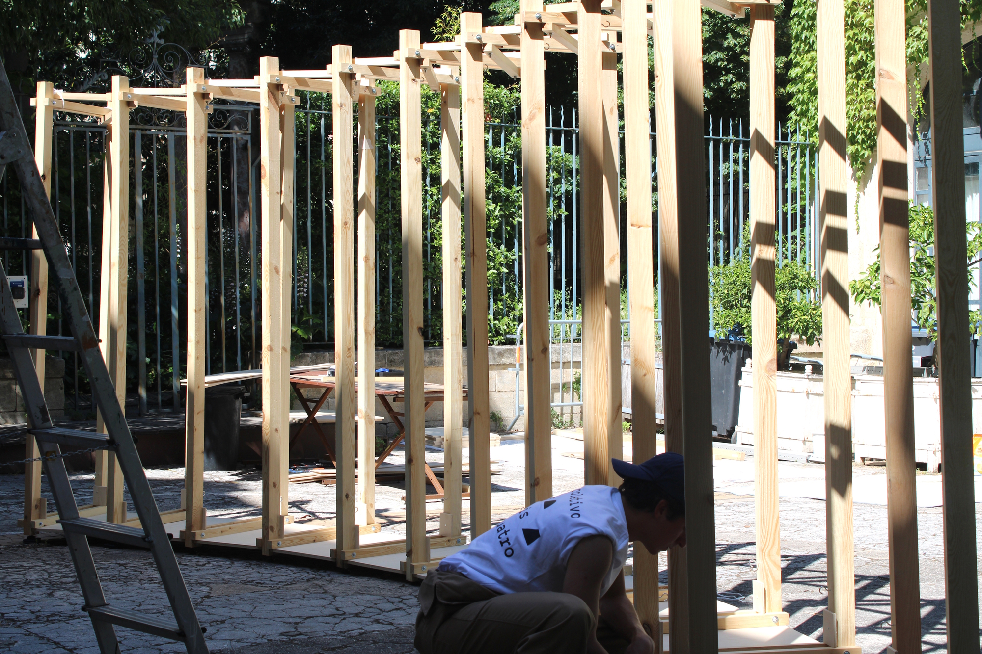 Row of bare timber posts in dappled sunlight with a crouching worker, a ladder and an iron fence behind