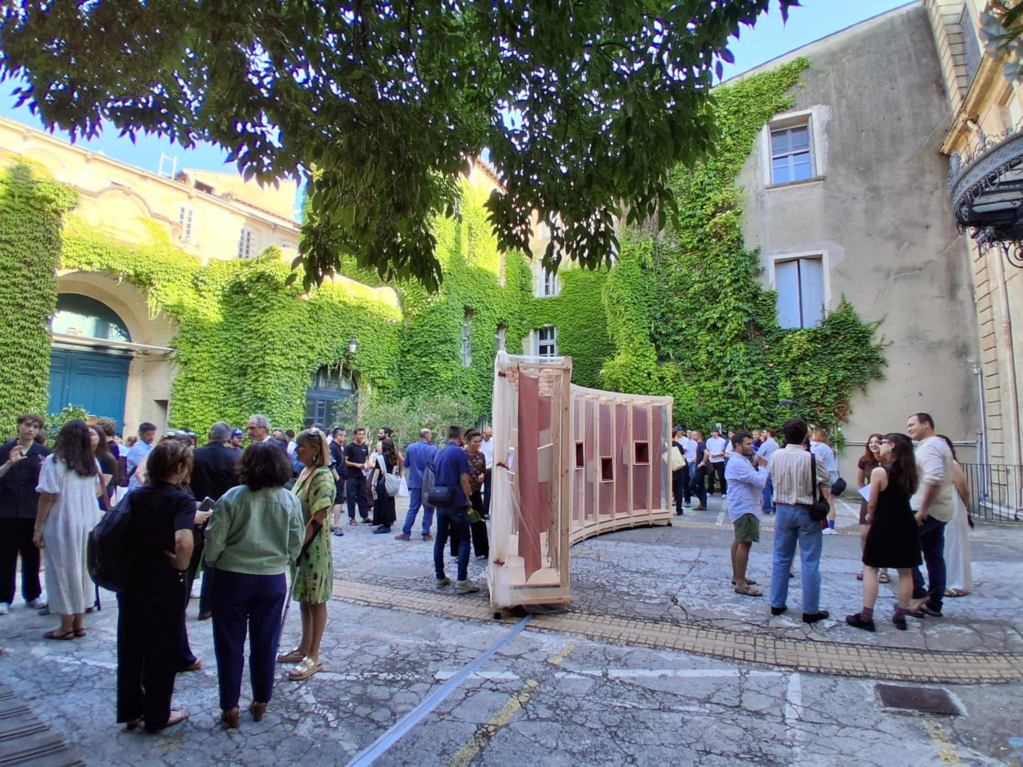 Curved timber pavilion with red fabric panels in a courtyard where a crowd gathers beneath ivy-covered walls and tree branches