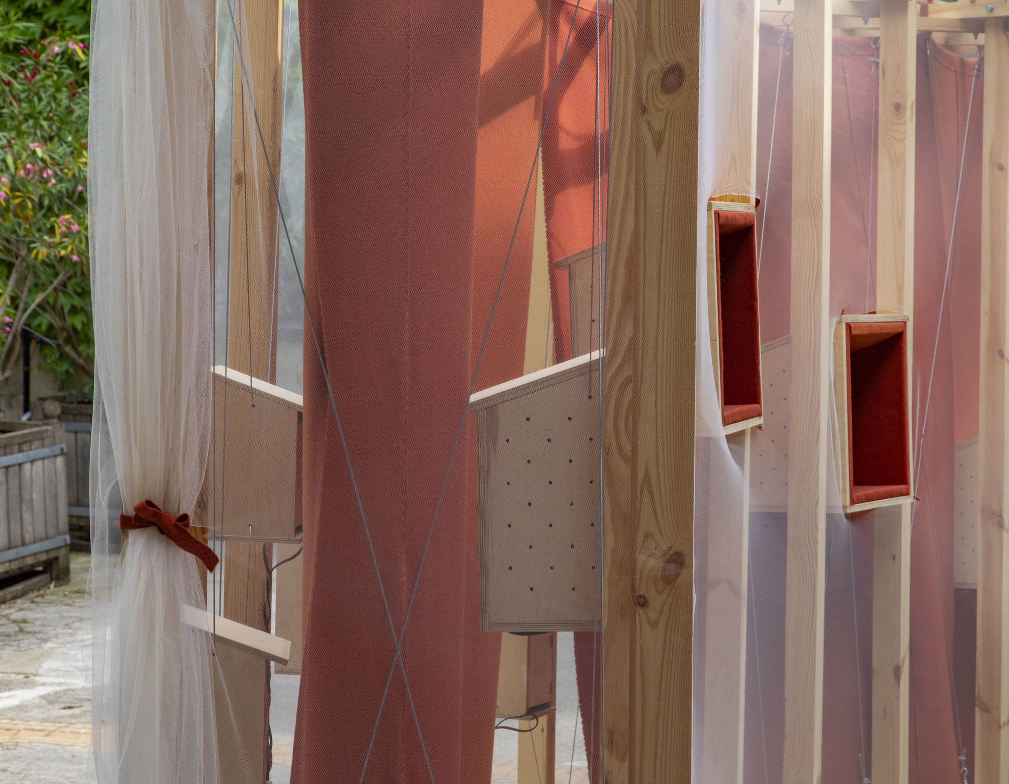 Close-up of pine posts, sheer tied curtains and rust-red fabric with small plywood boxes, seen against a flowering shrub