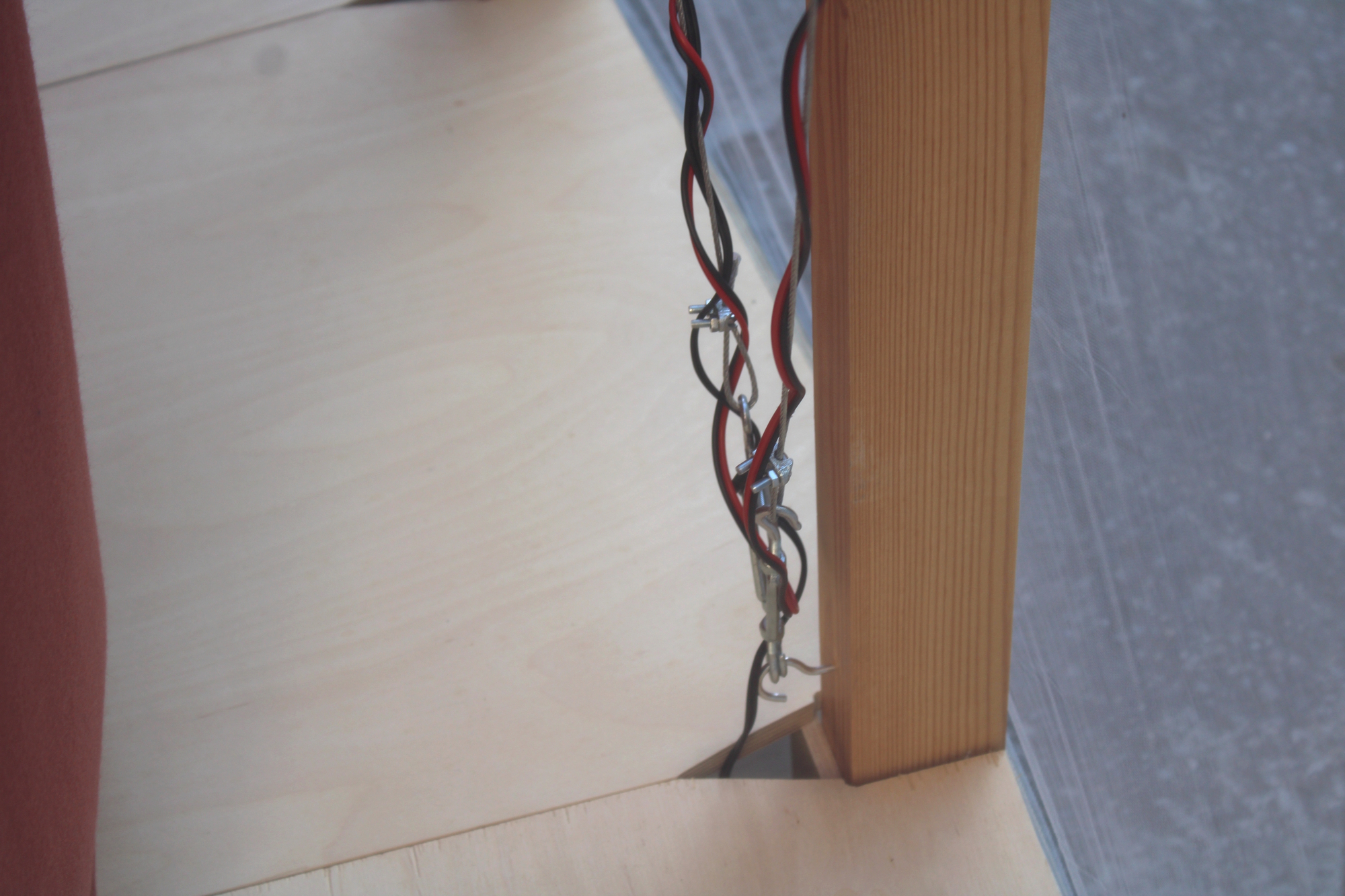 Detail of twisted red and black wires clipped beside a pine post at a birch plywood corner, behind translucent plastic sheeting