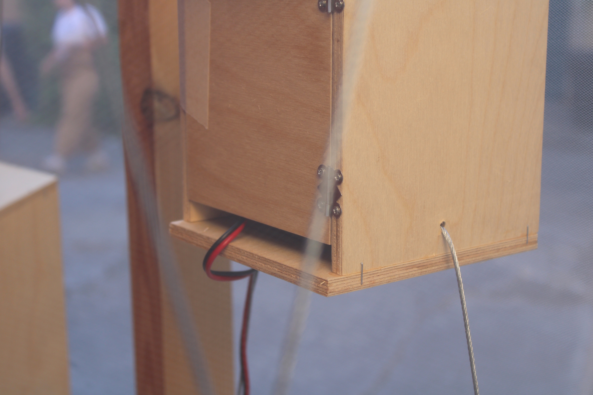 Close-up of a plywood box with small hinges, a red and black cable and a steel cord, with a blurred passerby behind
