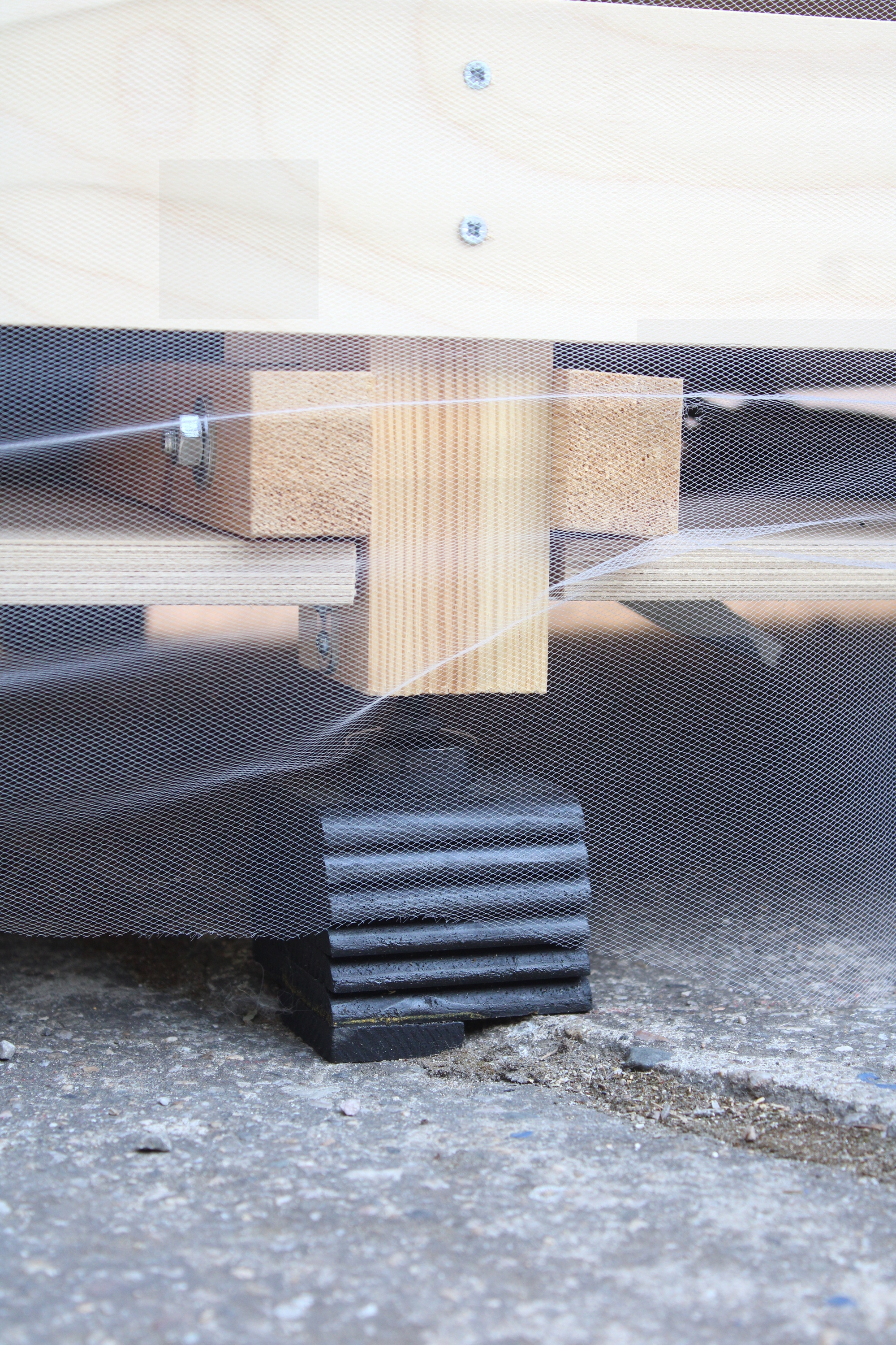 Close-up of a timber post resting on a black ribbed rubber foot over rough concrete, seen through fine mesh netting