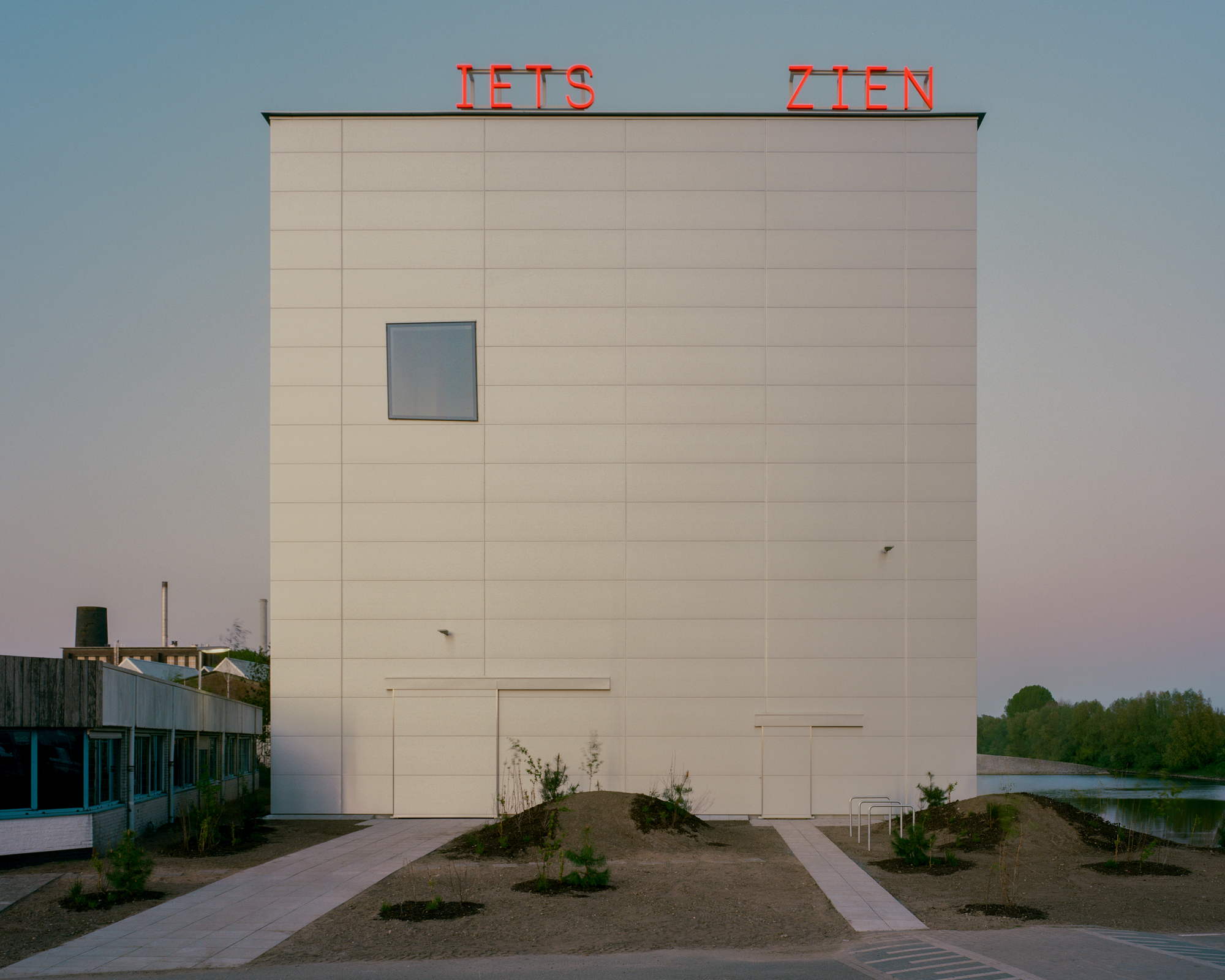 Front elevation of the white panelled cube with red neon lettering on its roof edge, seen at dusk beside a pond