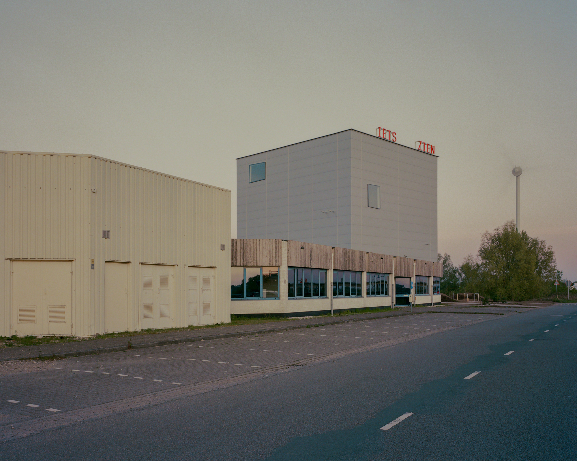 Corner view of the cube above a low timber-clad wing, with a wind turbine rising behind in the evening sky