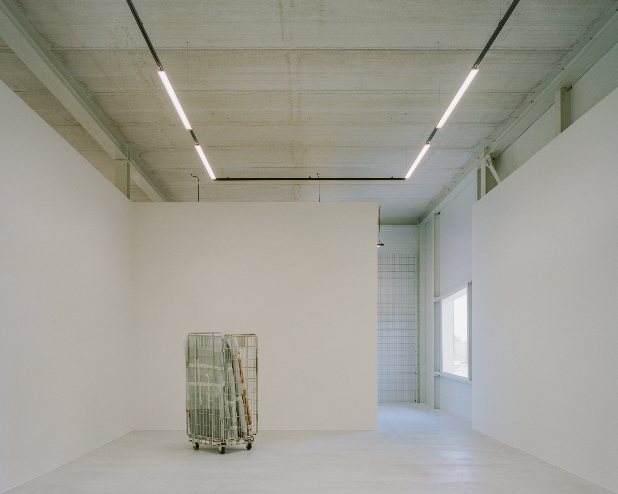 Gallery room with freestanding white walls, track lights and a metal roll cage on a pale concrete floor