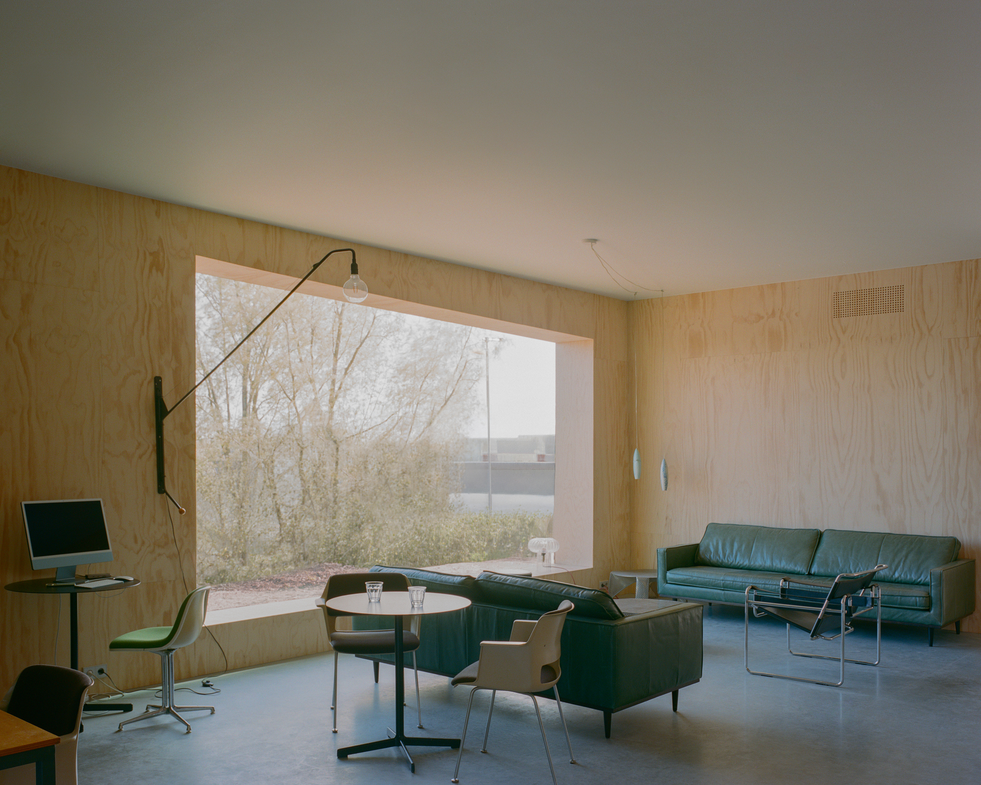 Plywood-lined lounge with a large framed window onto bare trees, green leather sofas and a long-armed wall lamp