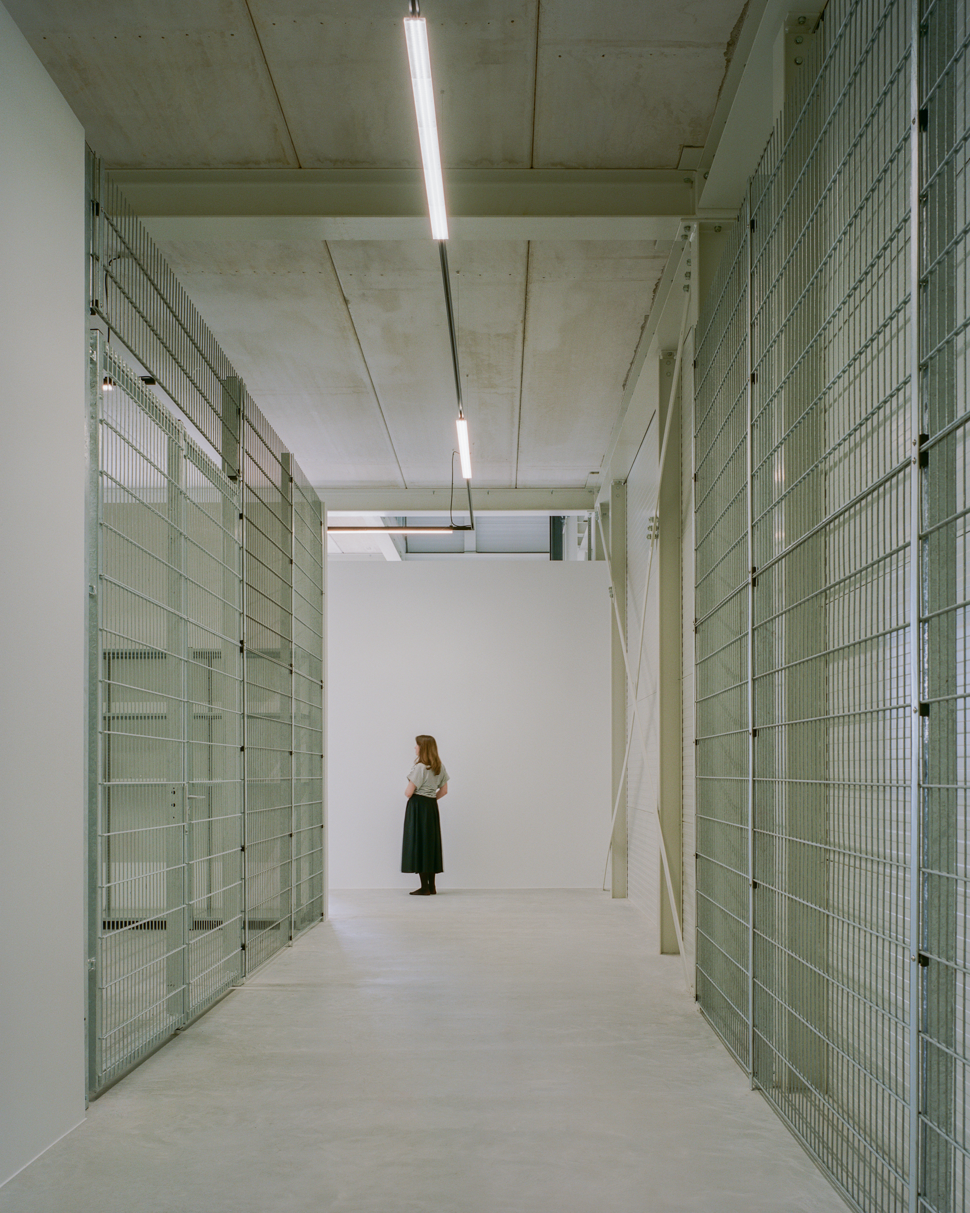 Corridor lined with galvanised mesh partitions and a woman in a dark skirt standing at the far white wall