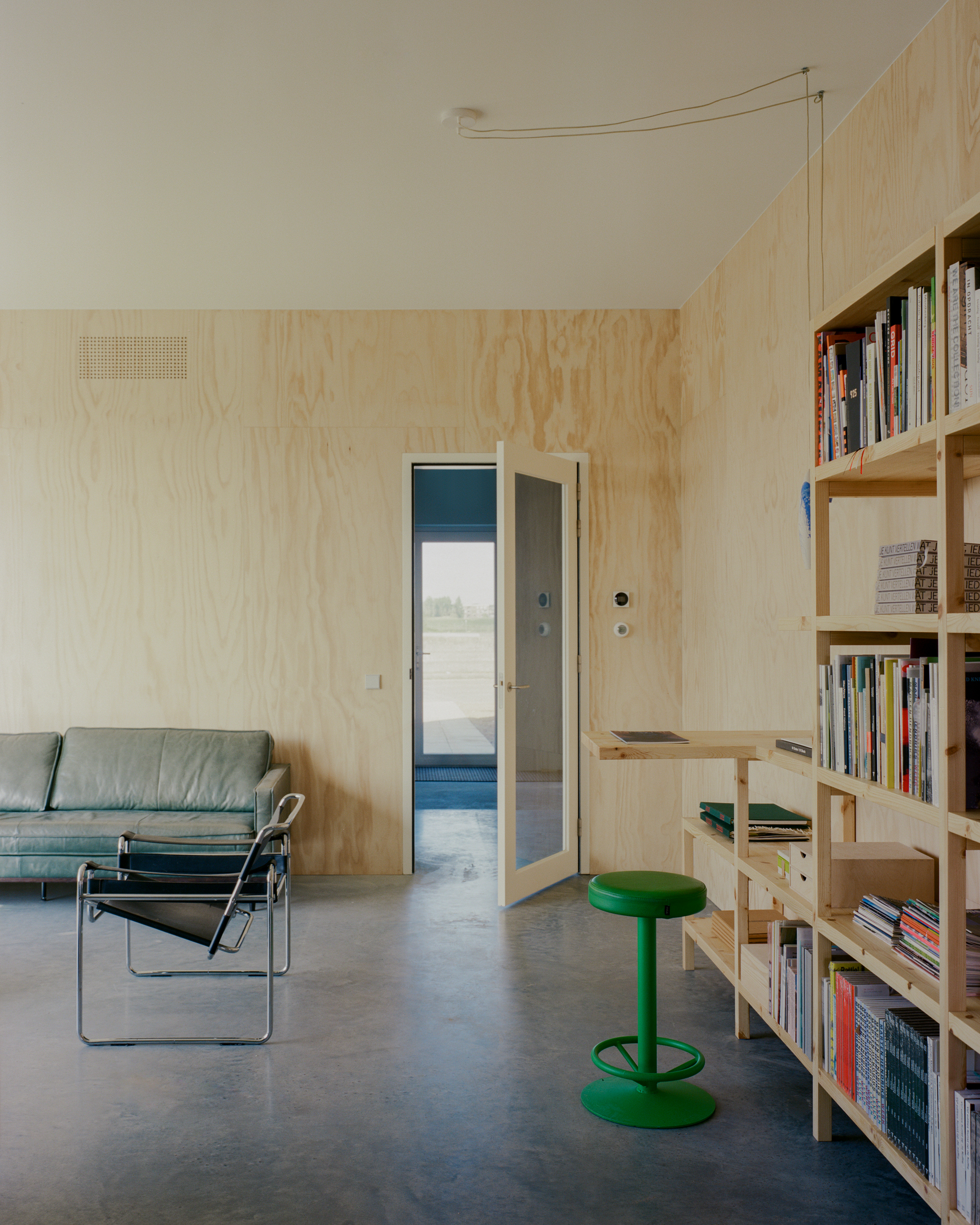 Plywood-lined room with a green leather sofa, steel-framed chair, green stool and timber bookshelf beside an open glazed door