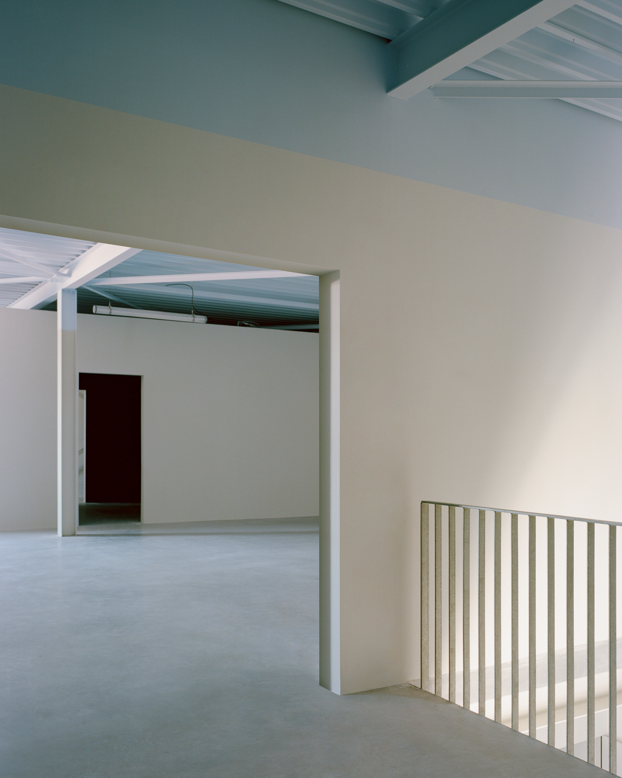 Upper-floor interior with a deep wall opening onto a steel-beamed room, and a slatted balustrade at the stair edge in cool daylight