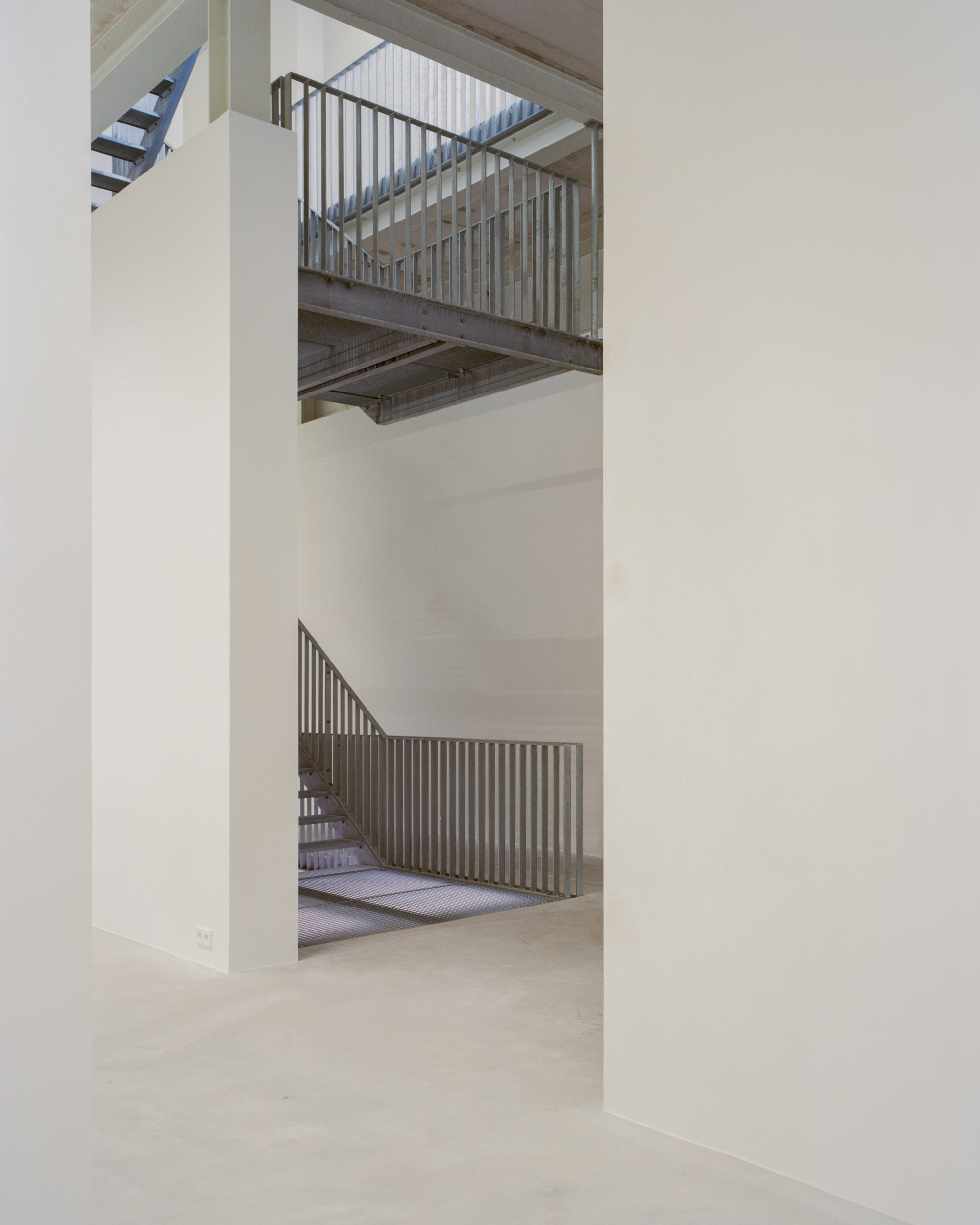 White walls frame a steel stair with slatted metal balustrade and a catwalk above, over a pale concrete floor