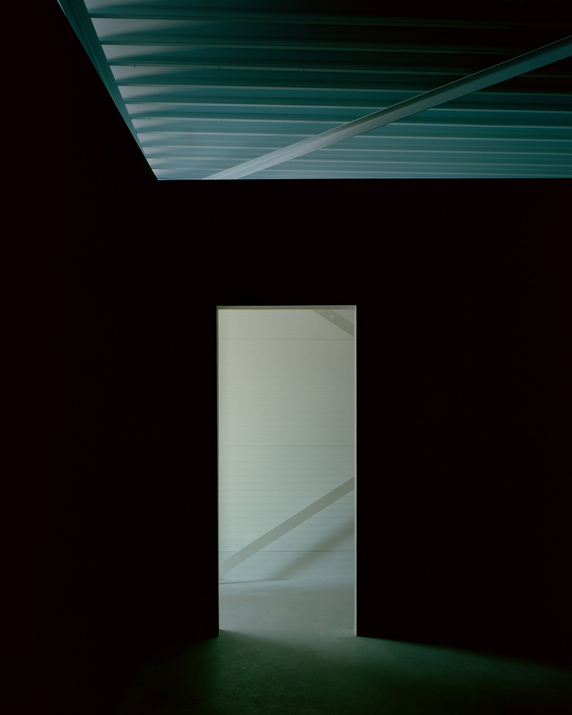Dark room with a lit doorway opening onto a white space, corrugated ceiling panel glowing green overhead