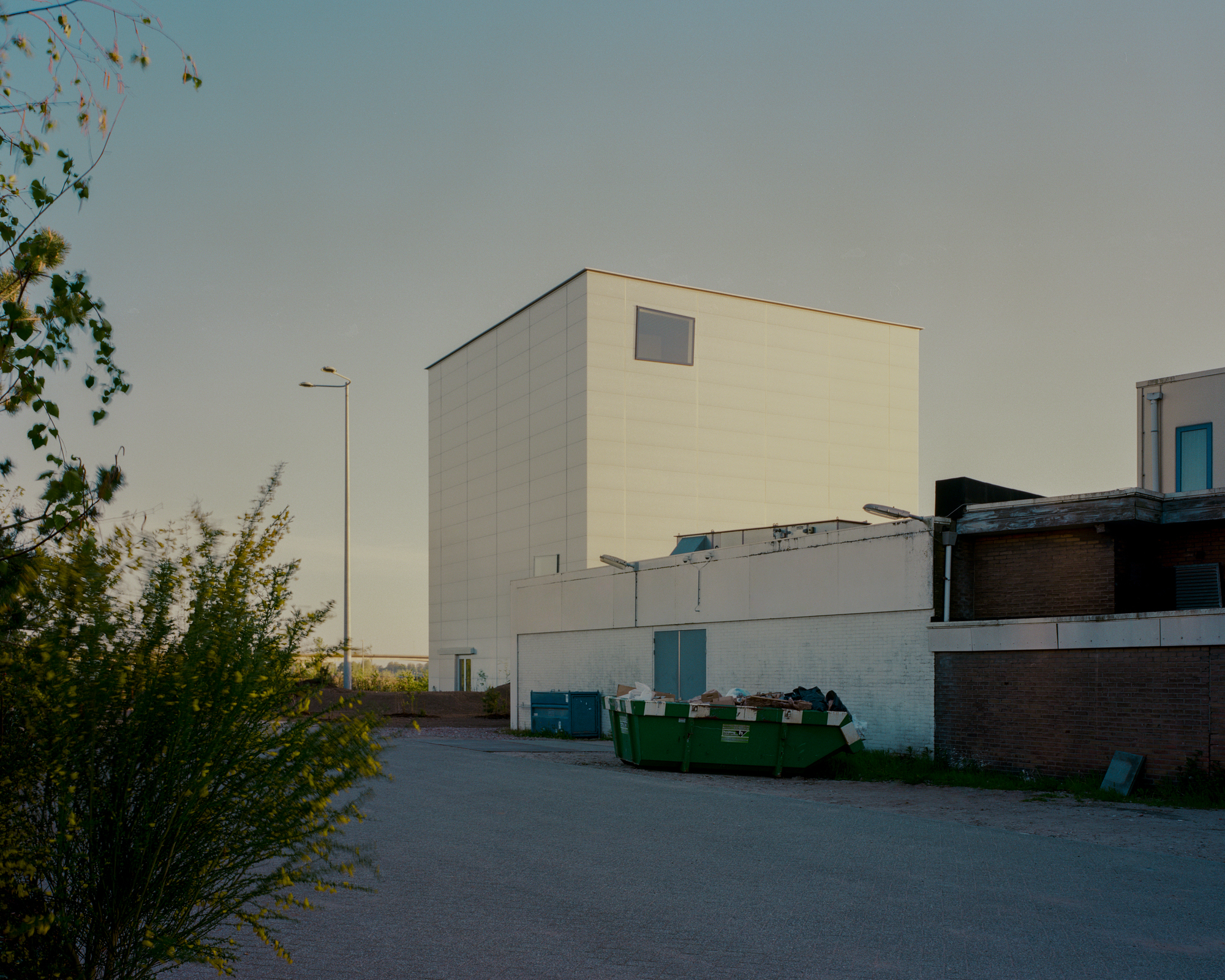 Pale cubic volume with a single window rising behind a green skip and brick outbuildings in low evening light