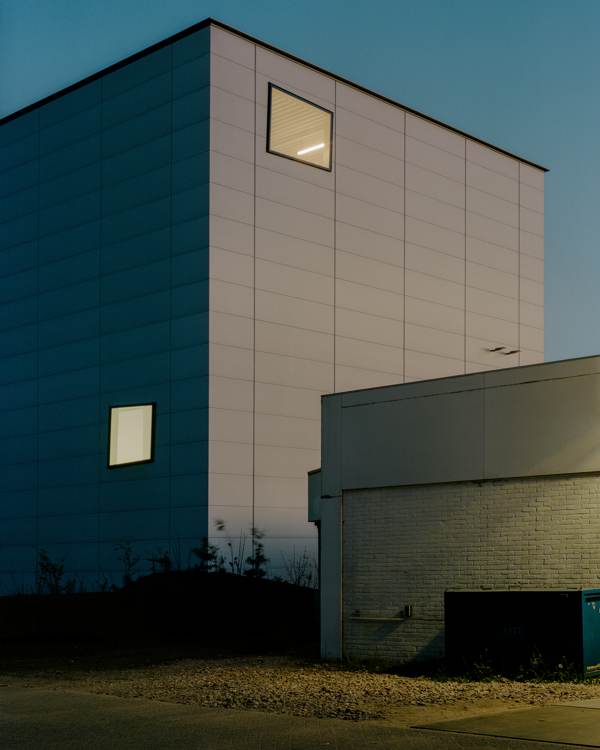 Panelled cube facade with two lit windows against a deep blue dusk sky beside a white brick wall
