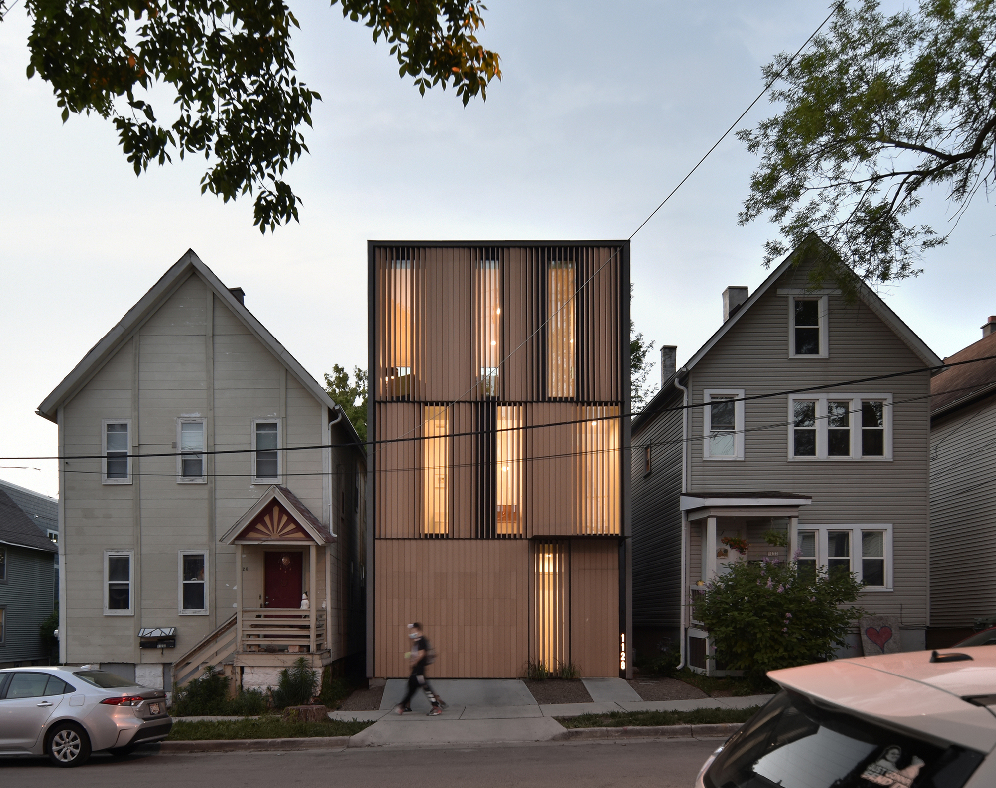 Narrow timber-louvered house glowing between two gabled wood-frame neighbors at dusk, with a pedestrian walking past on the sidewalk