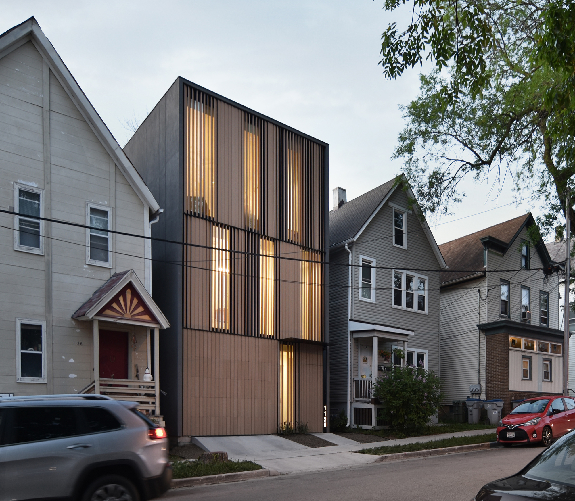 Angled street view of the slatted wood and dark metal facade lit from within, flanked by older houses under an evening sky