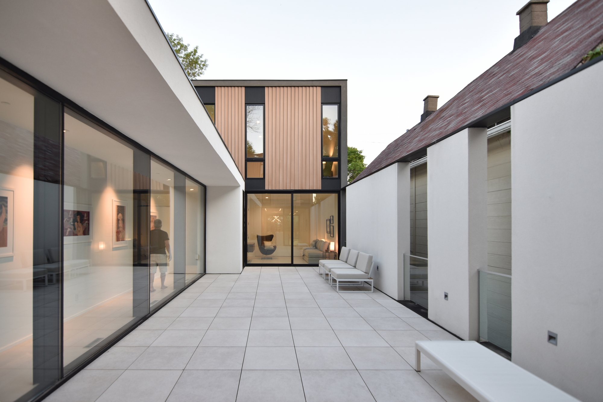 Paved courtyard framed by a glazed gallery wing and white walls, with a timber-clad upper volume beyond at dusk