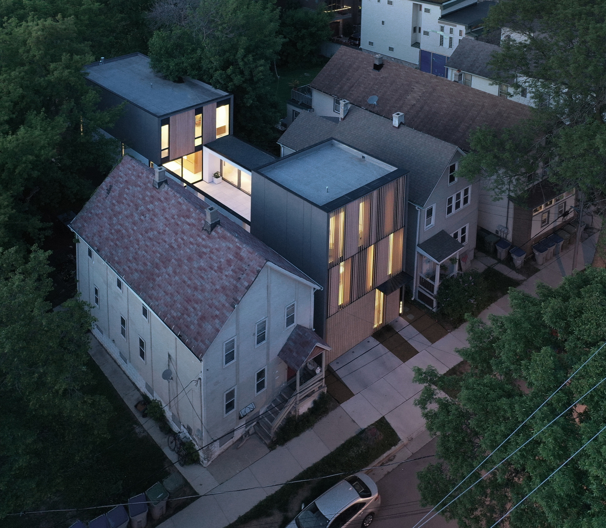 Aerial view of the flat-roofed louvered volume and rear courtyard wing squeezed between pitched-roof houses among dark trees at dusk