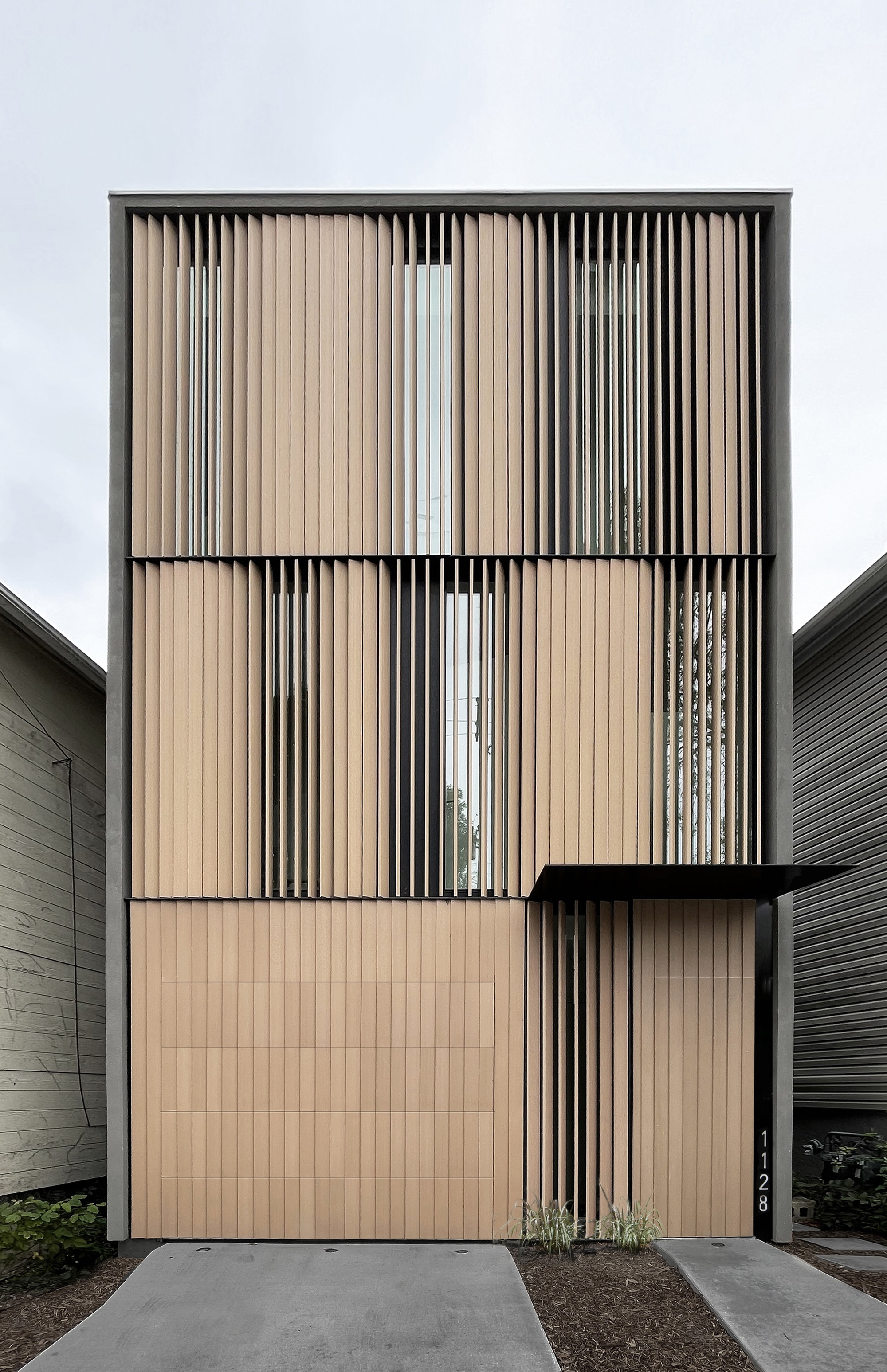 Front elevation of the three-storey facade with vertical wood louvers, tall slot windows and a cantilevered canopy over the entry, under overcast sky