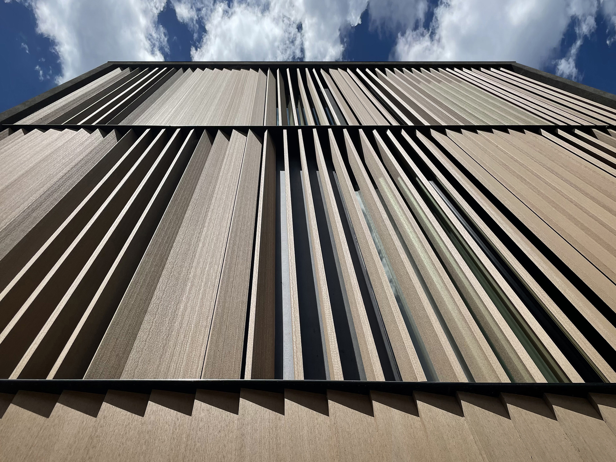 Low upward view of angled vertical timber fins over glazing, set against blue sky and scattered white clouds