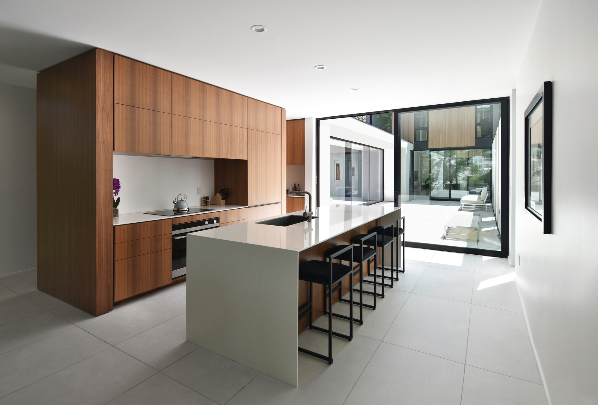 Kitchen with walnut cabinetry and a white island with black stools, opening through sliding glass to a sunlit courtyard