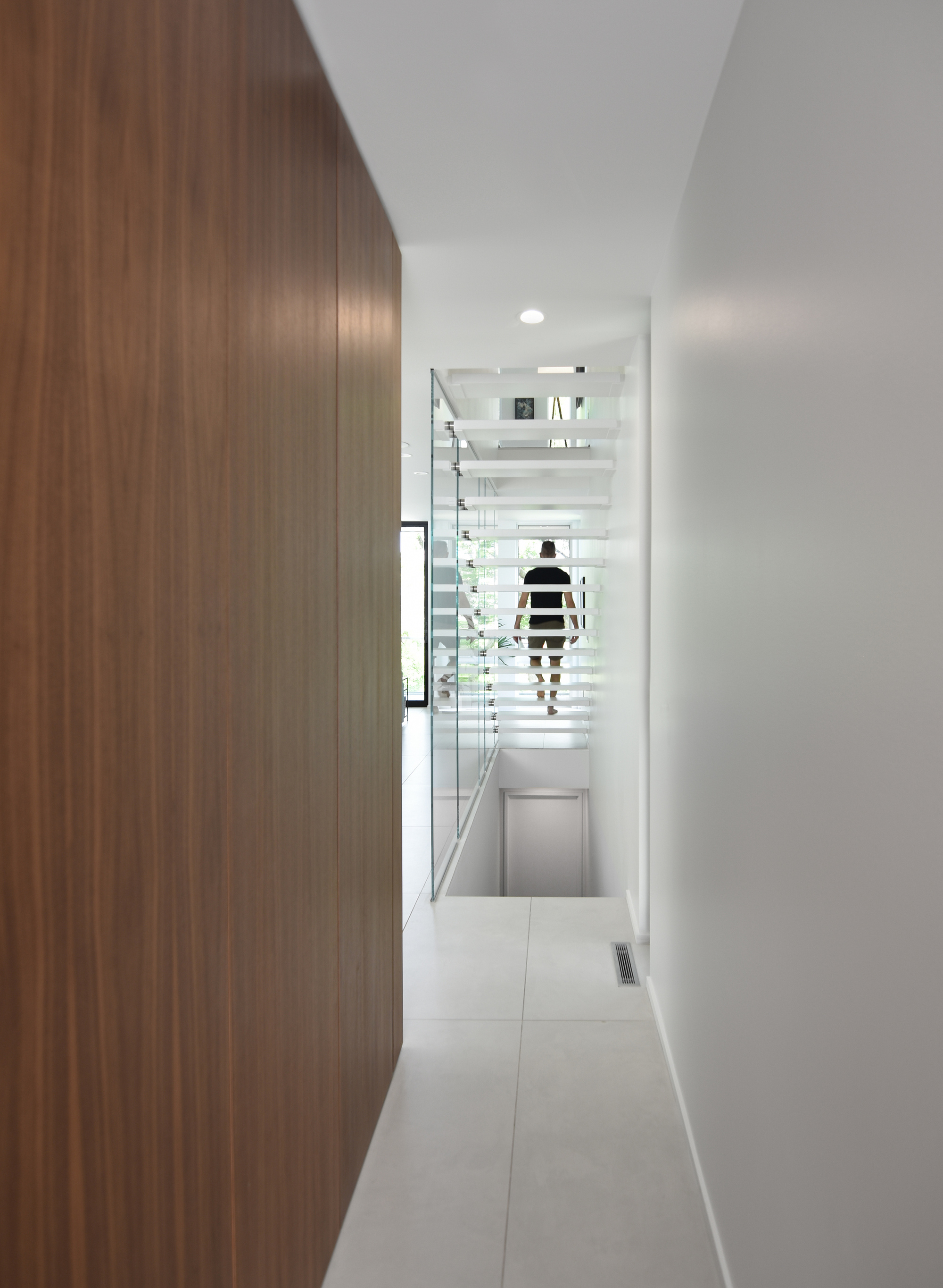 Narrow hallway beside a walnut panel wall leading to a glass-sided floating stair, with a person climbing in the distance