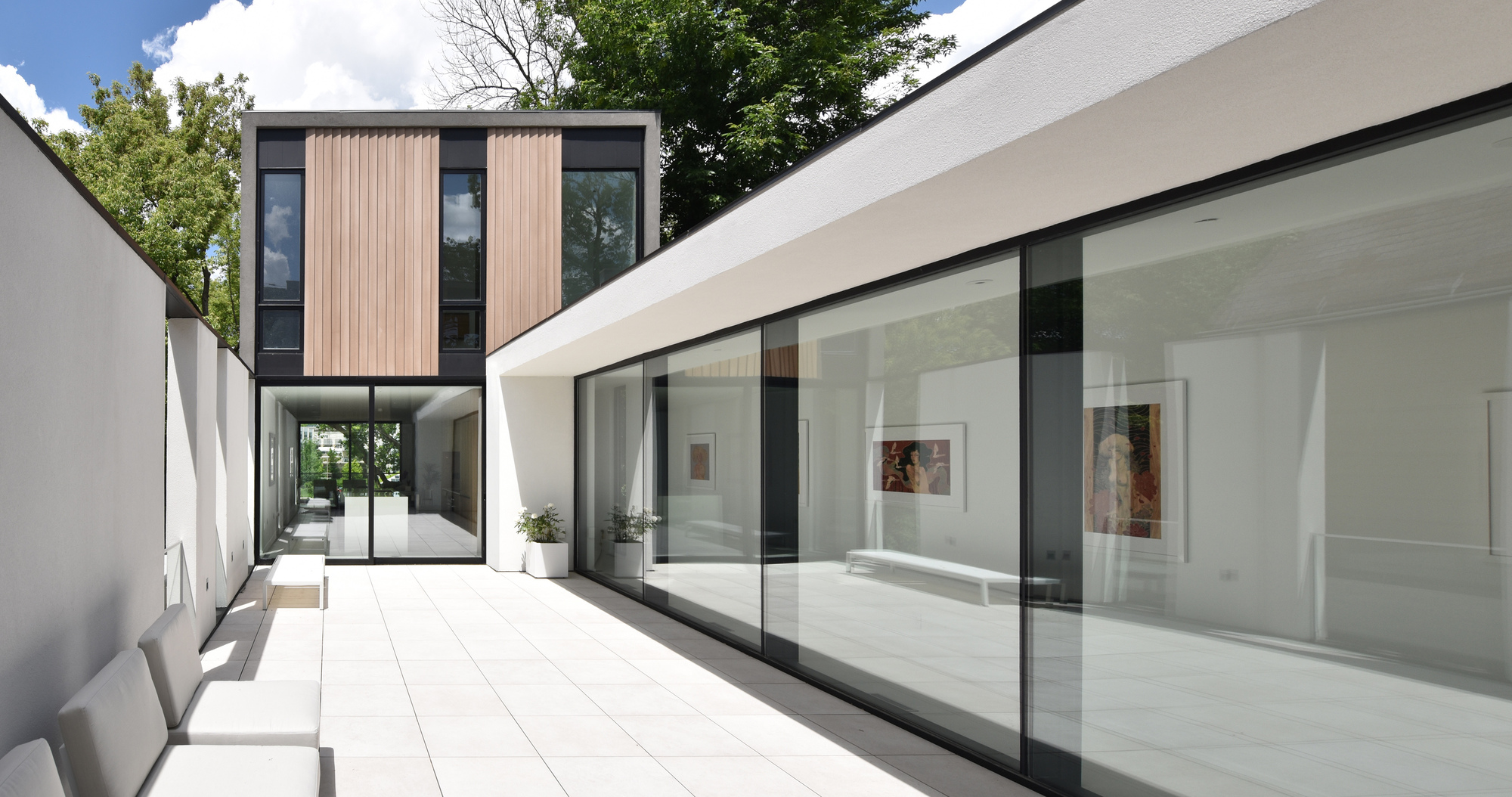 Courtyard terrace with white paving, long sliding glass wall and a timber-clad upper volume under a bright summer sky with clouds
